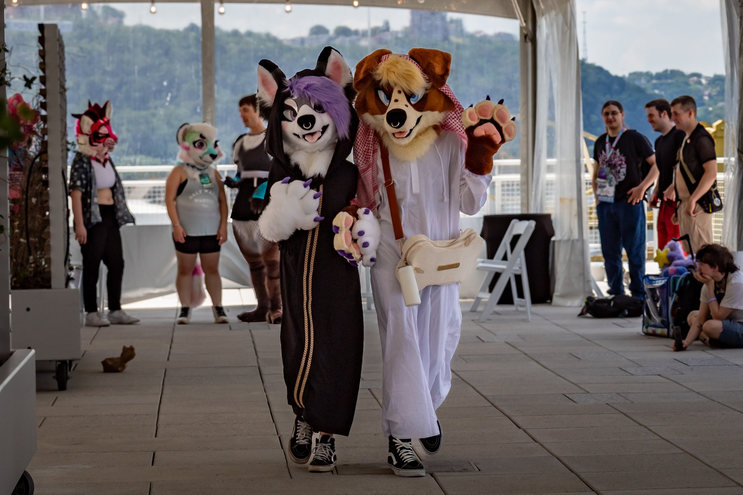Two people dressed in furry animal costumes, one as a black and white wolf and the other as a brown and beige dog, walking inside a tent with other people in the background. The wolf costume has purple accents and the dog costume is wearing a traditi