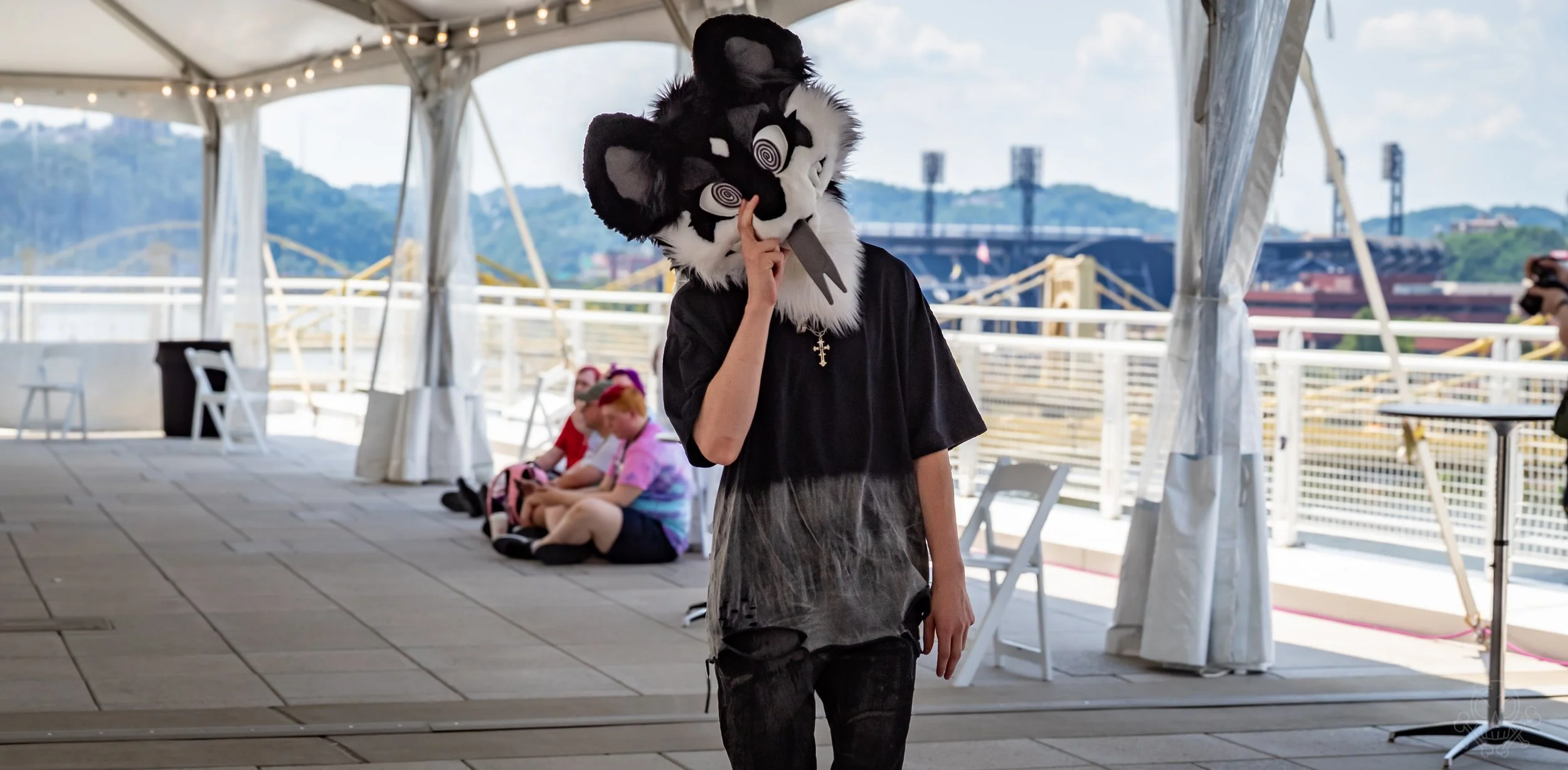Person wearing a black and white cat mask, black T-shirt, and ripped jeans standing under a tent on a balcony with a mountain view.