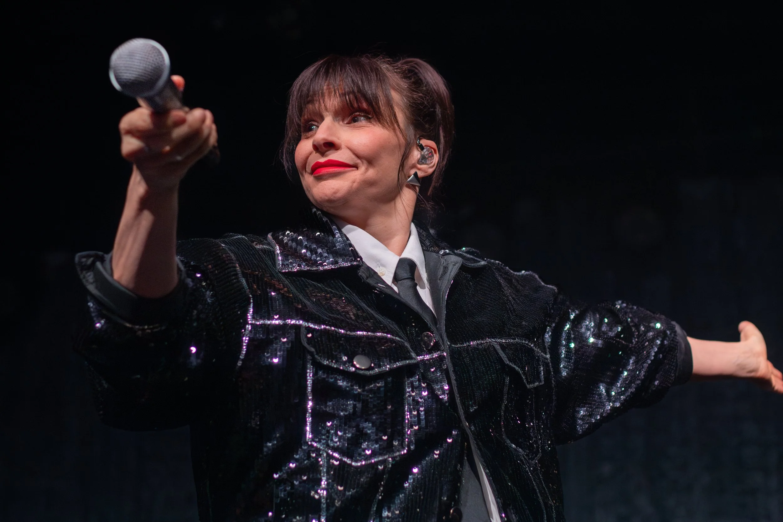 A woman holding a microphone on stage, wearing a shiny black jacket and a white shirt with a tie, with her arms outstretched.