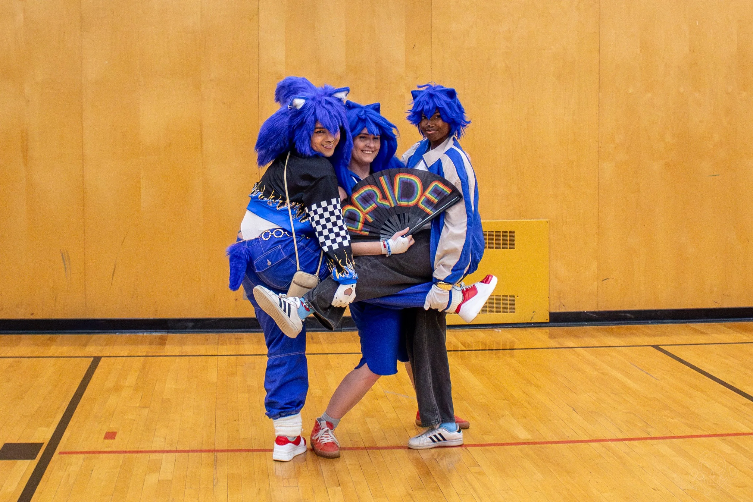 Three people in blue furry lion costumes with tails, two of them carrying someone dressed in colorful athletic clothing holding a rainbow-colored fan with the word 'PRIDE' on it, in a gymnasium.
