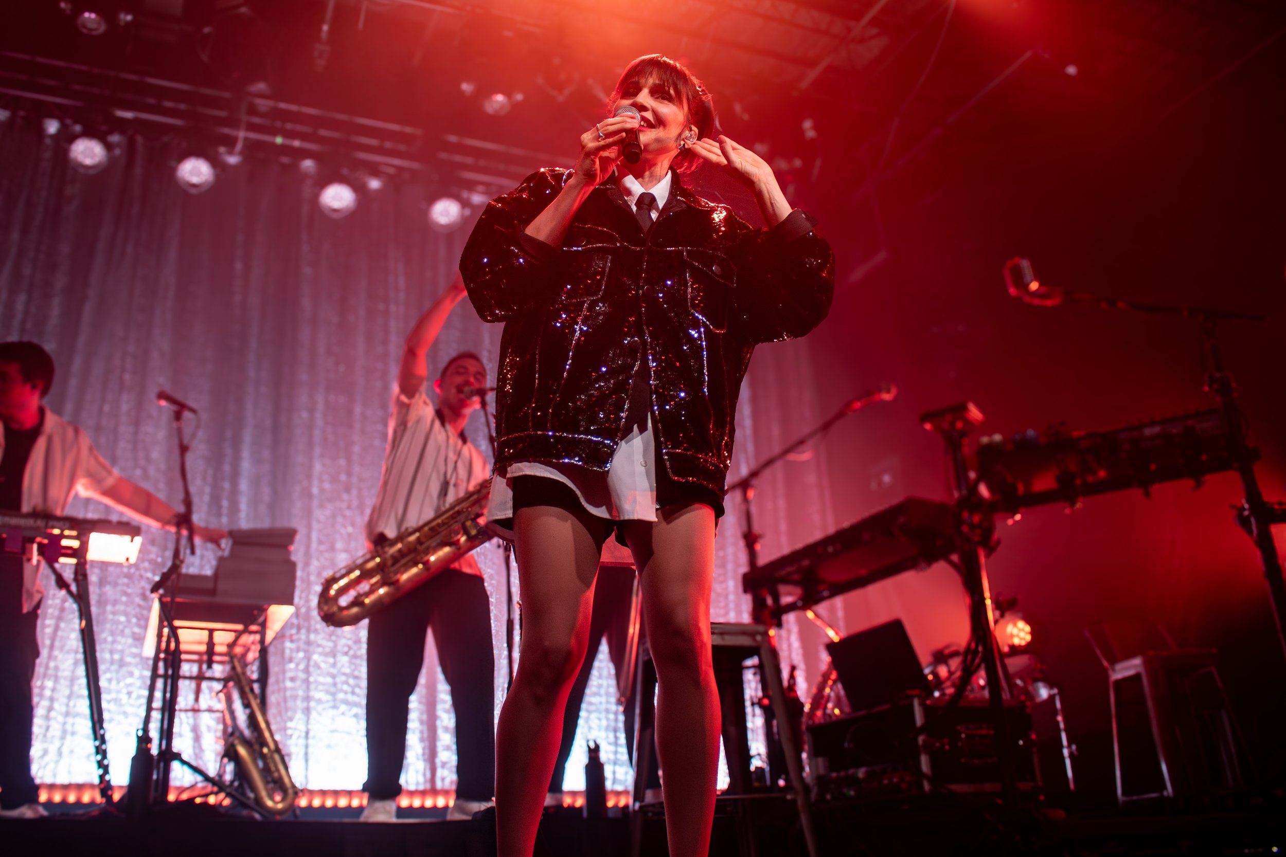 A female singer performing on stage with backup musicians, surrounded by musical equipment and red stage lighting.