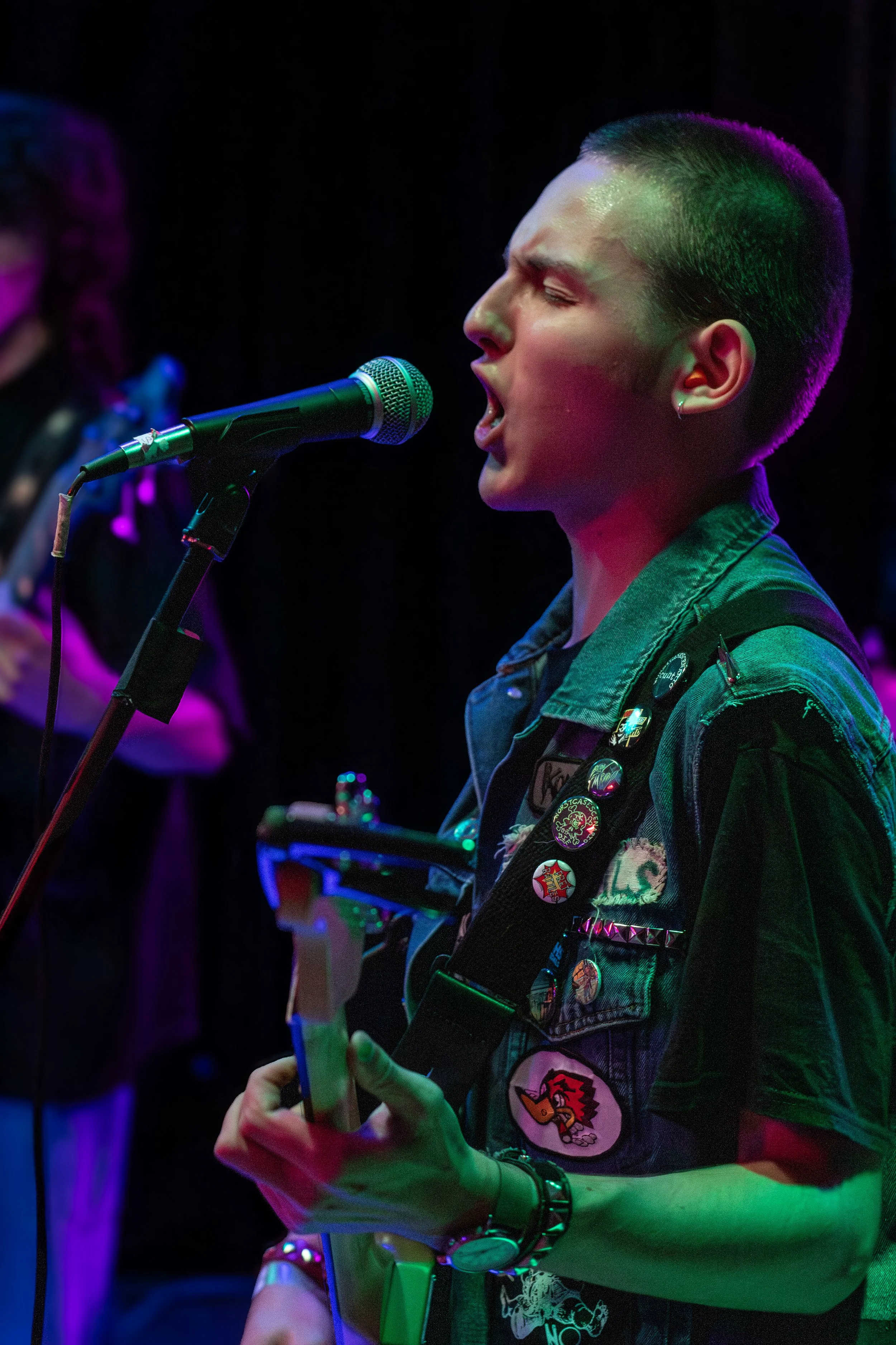 A person singing into a microphone while playing an electric guitar, wearing a denim vest with various pins and patches, in a dimly lit setting with colored stage lighting.