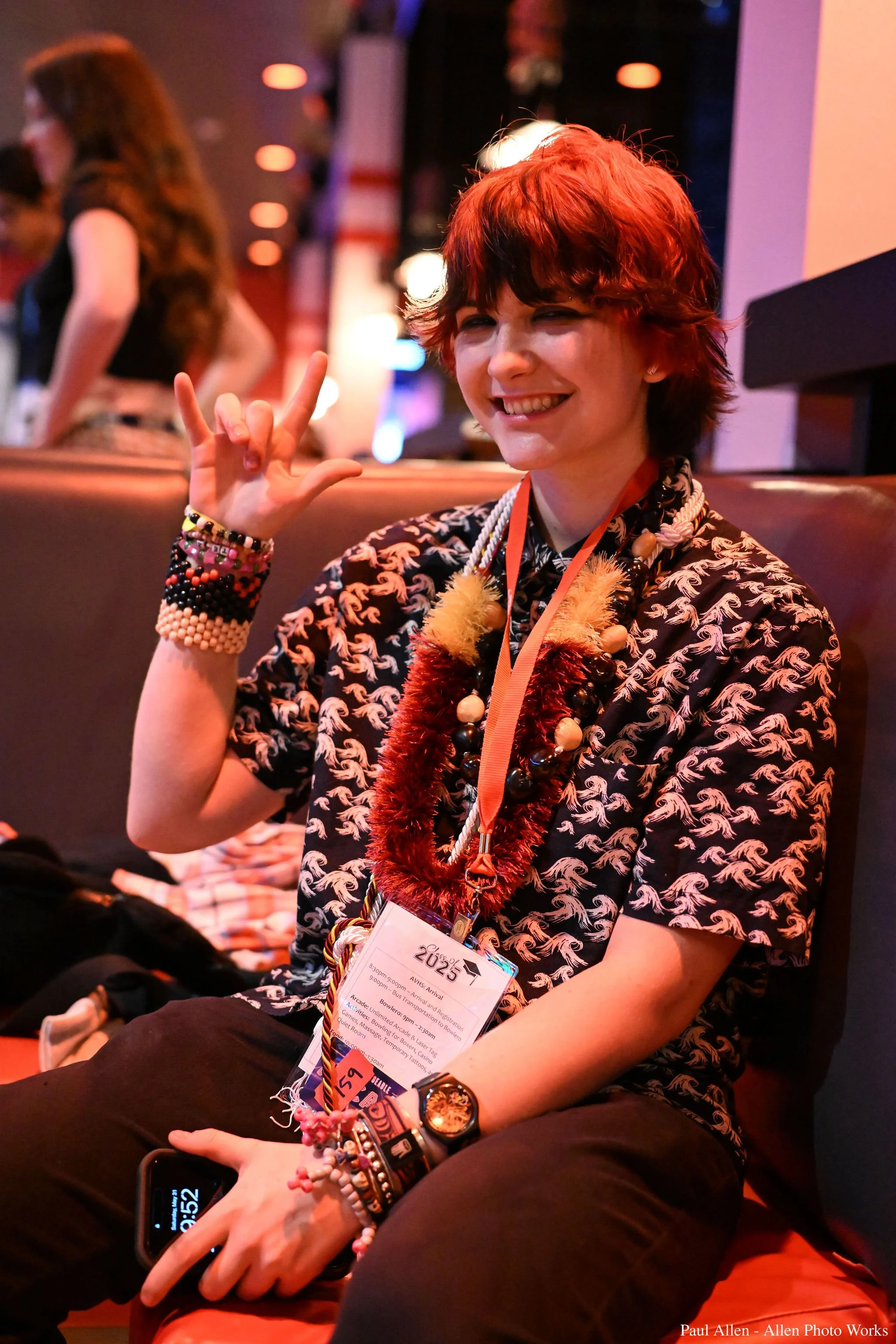 A person with short red hair smiling and flashing a peace sign, wearing a black patterned shirt and various colorful accessories, sitting in a dimly lit casual setting.