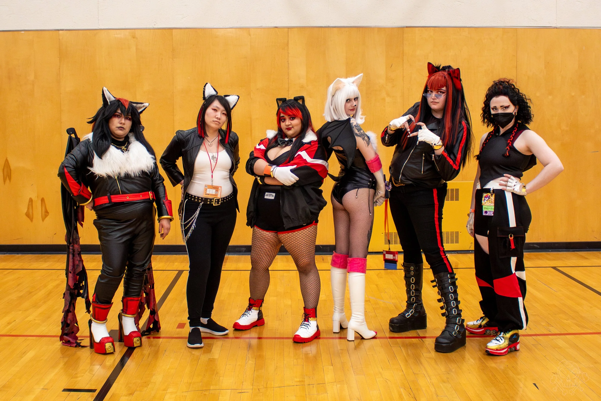 Six women dressed in cosplay costumes with cat-themed accessories, standing in a gymnasium with a wooden floor and yellow wall.