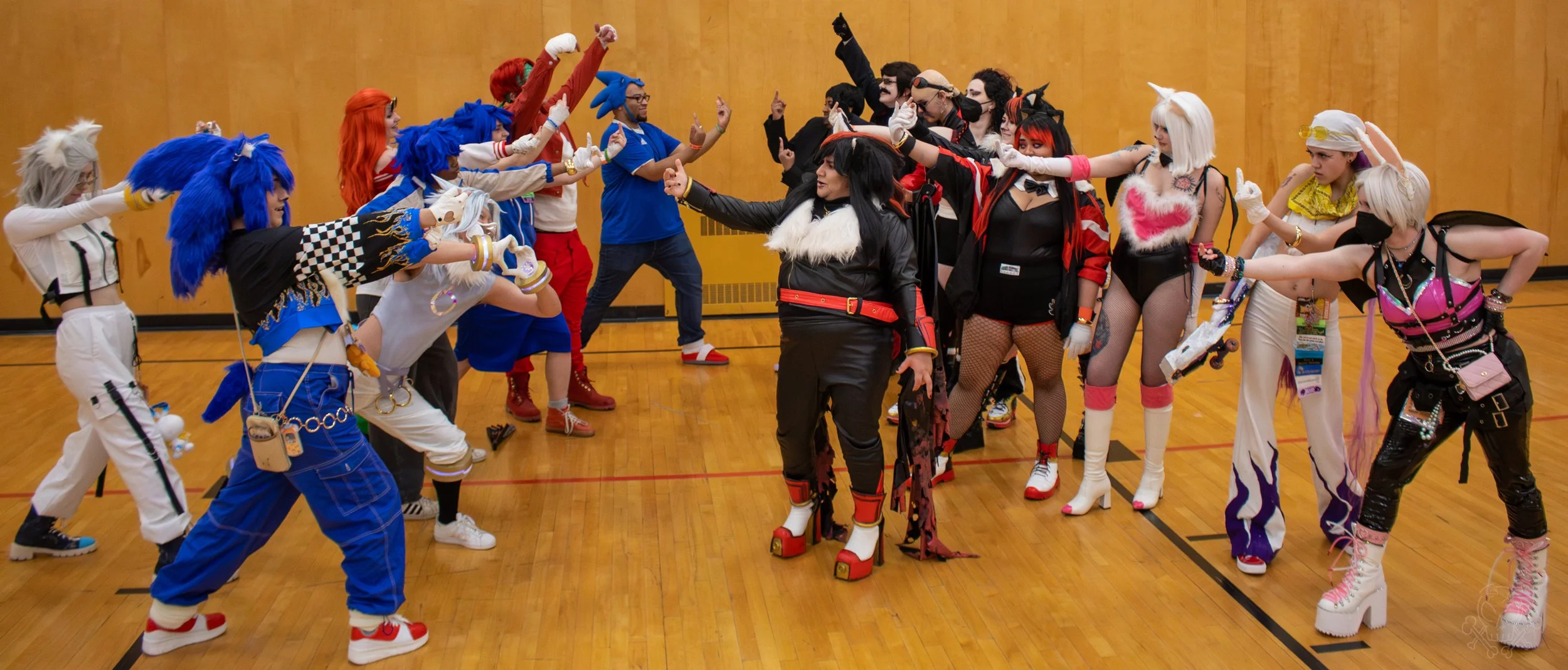 Group of people dressed in colorful, elaborate costumes facing each other in a gymnasium, seemingly participating in a costume contest or cosplay event.