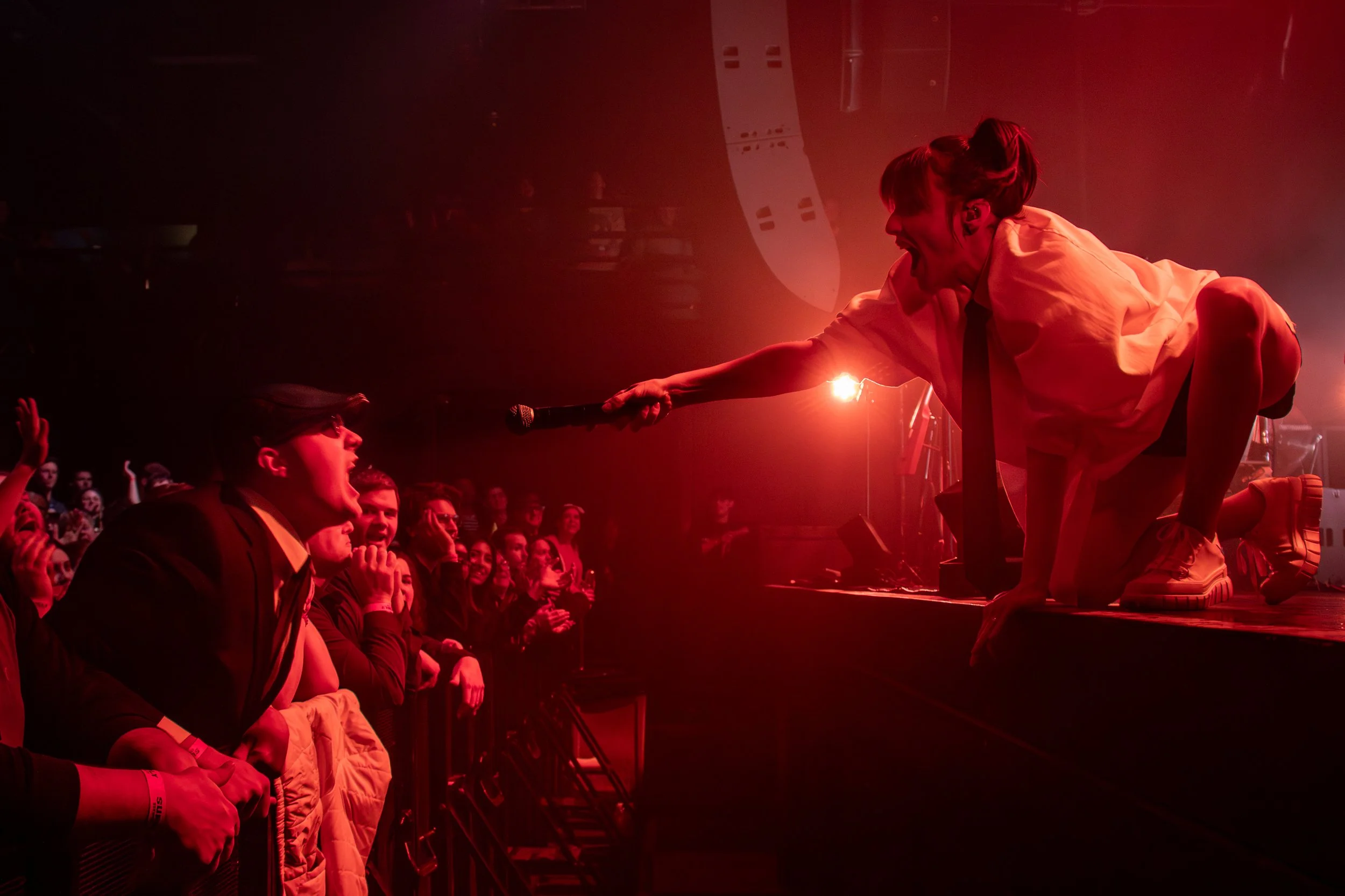 A female performer in a white shirt and black tie is crouching on stage, holding a microphone toward an audience member, who is wearing glasses and a black jacket, at a concert with red lighting.