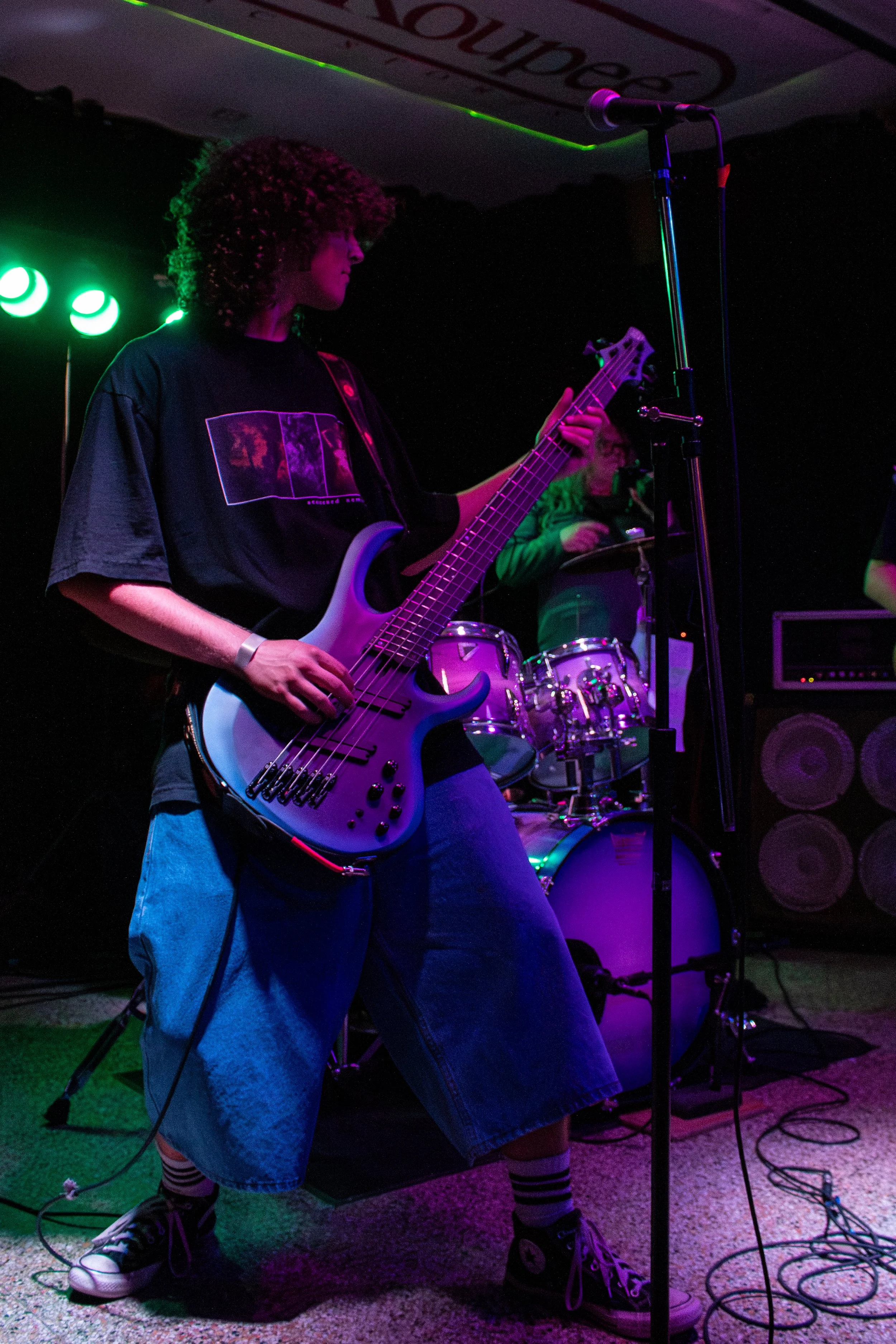 A young person with curly hair playing a bass guitar on stage, with a drummer in the background and colorful stage lighting.