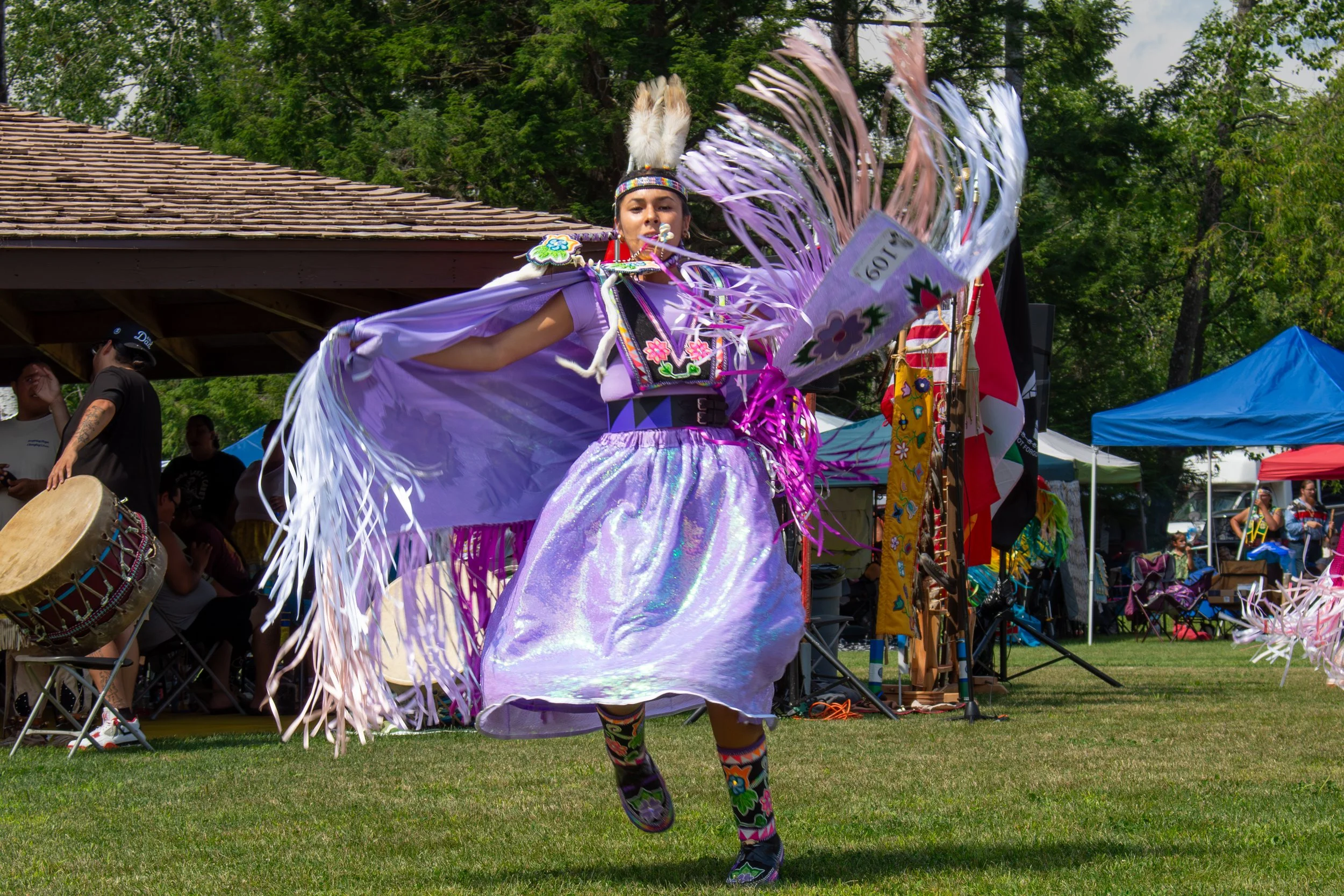 A woman in a vibrant purple traditional Native American dance costume with feathers, floral patterns, and iridescent fabric, performing a dance at an outdoor cultural event with tents and spectators in the background.