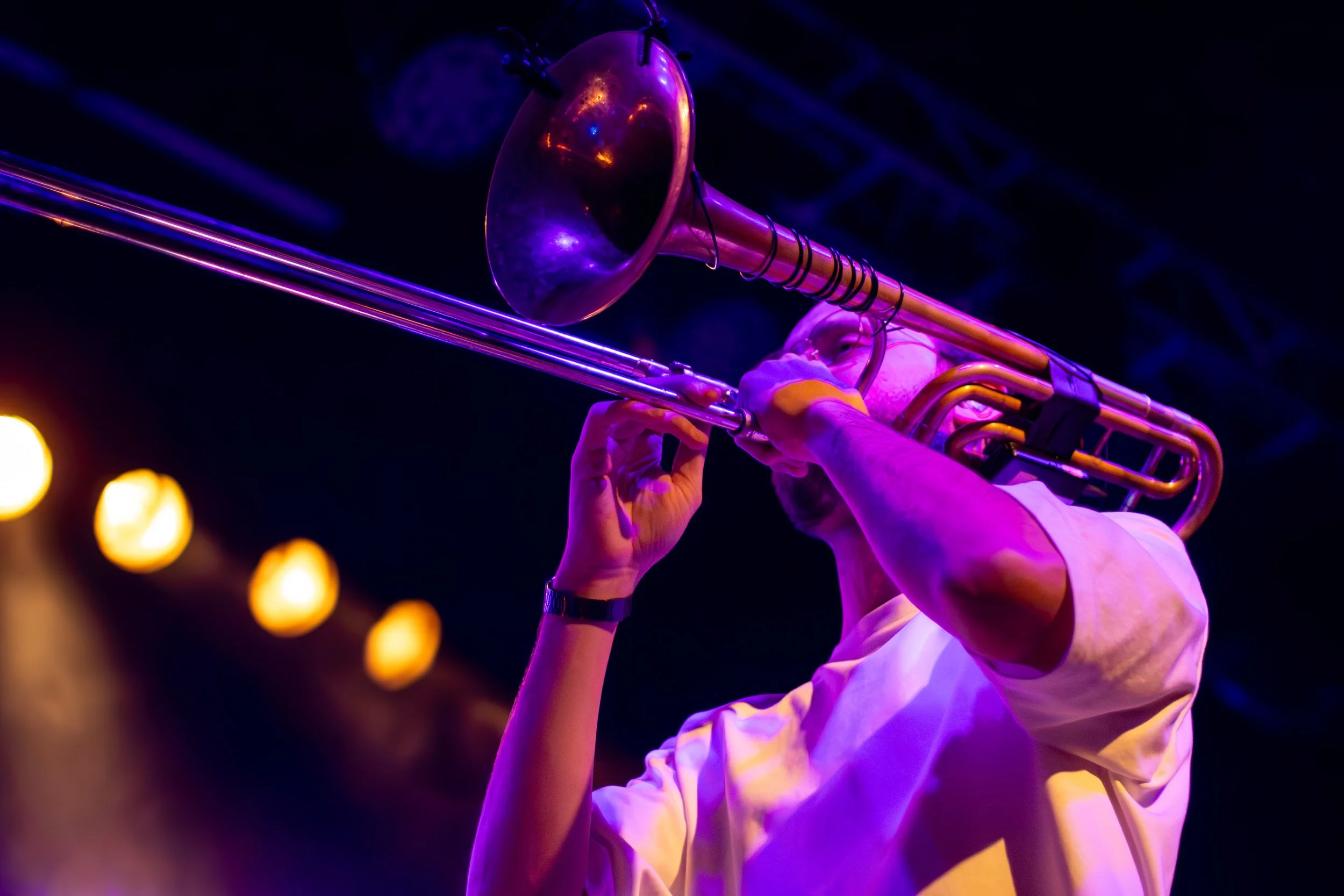 A performer playing a trombone on stage, illuminated by purple and yellow lighting.