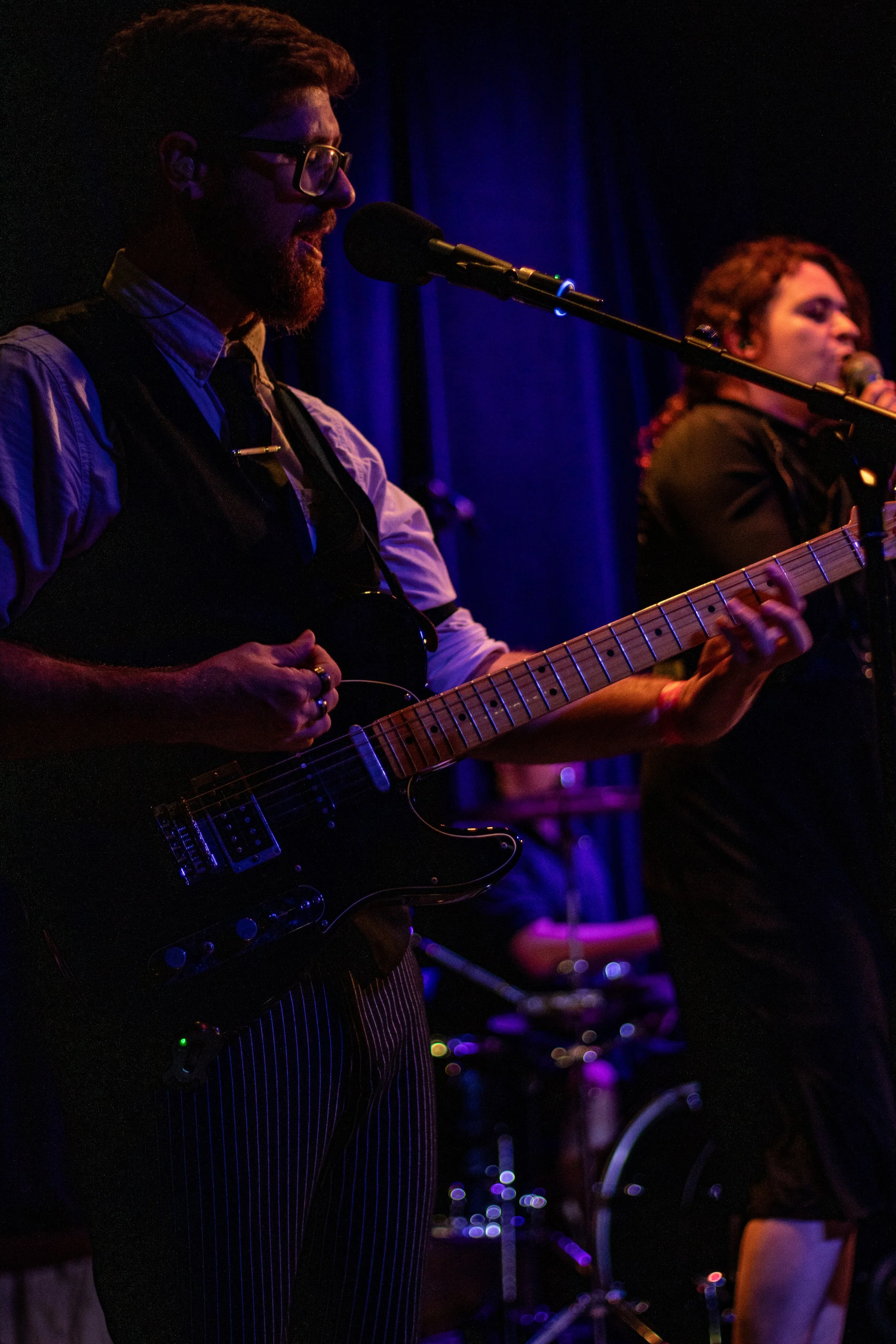 Two musicians performing on stage with microphones, one playing an electric guitar and the other singing into a microphone, with dim lighting and dark background.