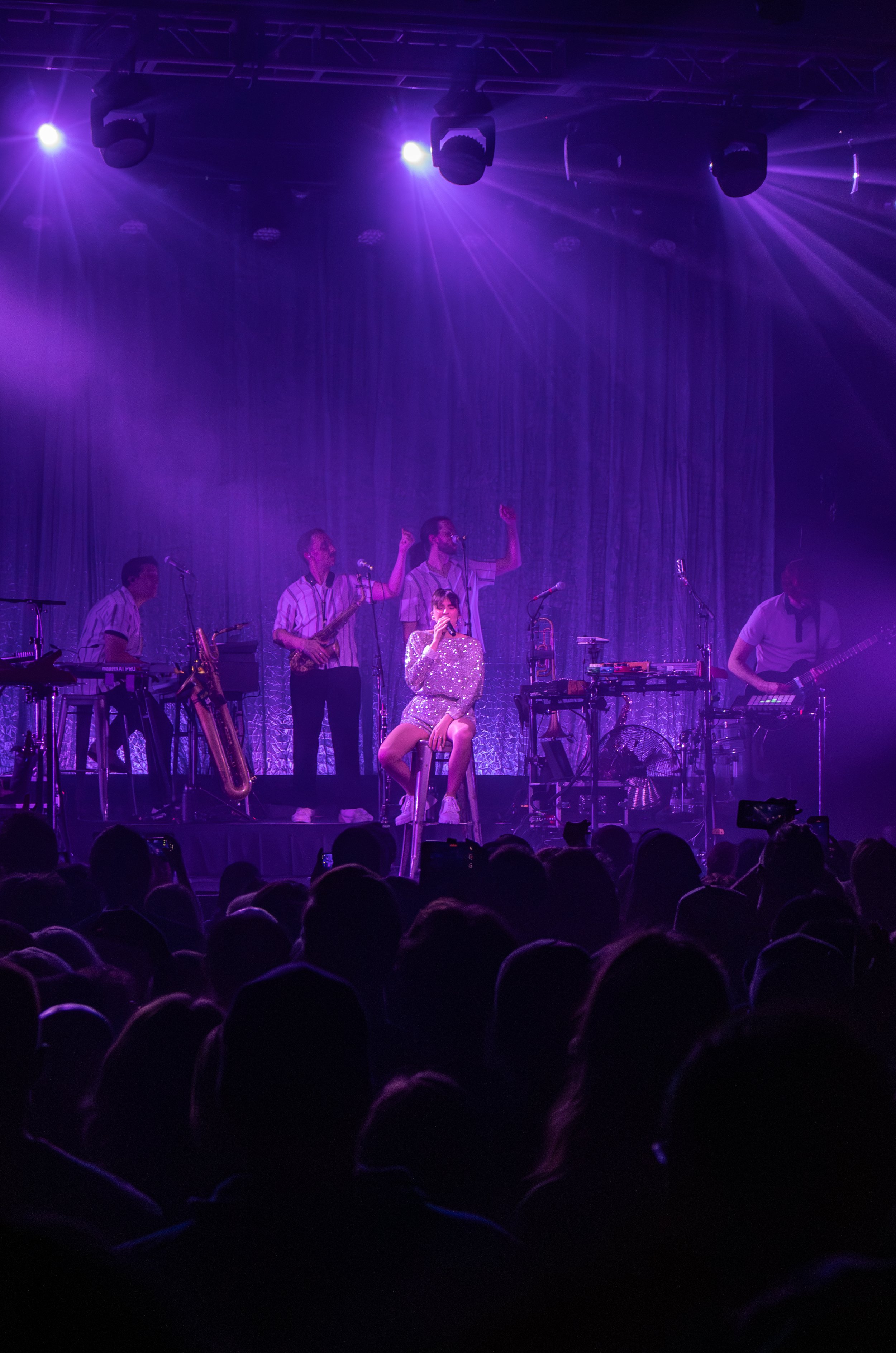 A live music performance on stage with a female singer sitting on a stool, surrounded by band members playing guitar, keyboard, drums, saxophone, and other instruments, under purple stage lighting, with an audience watching.