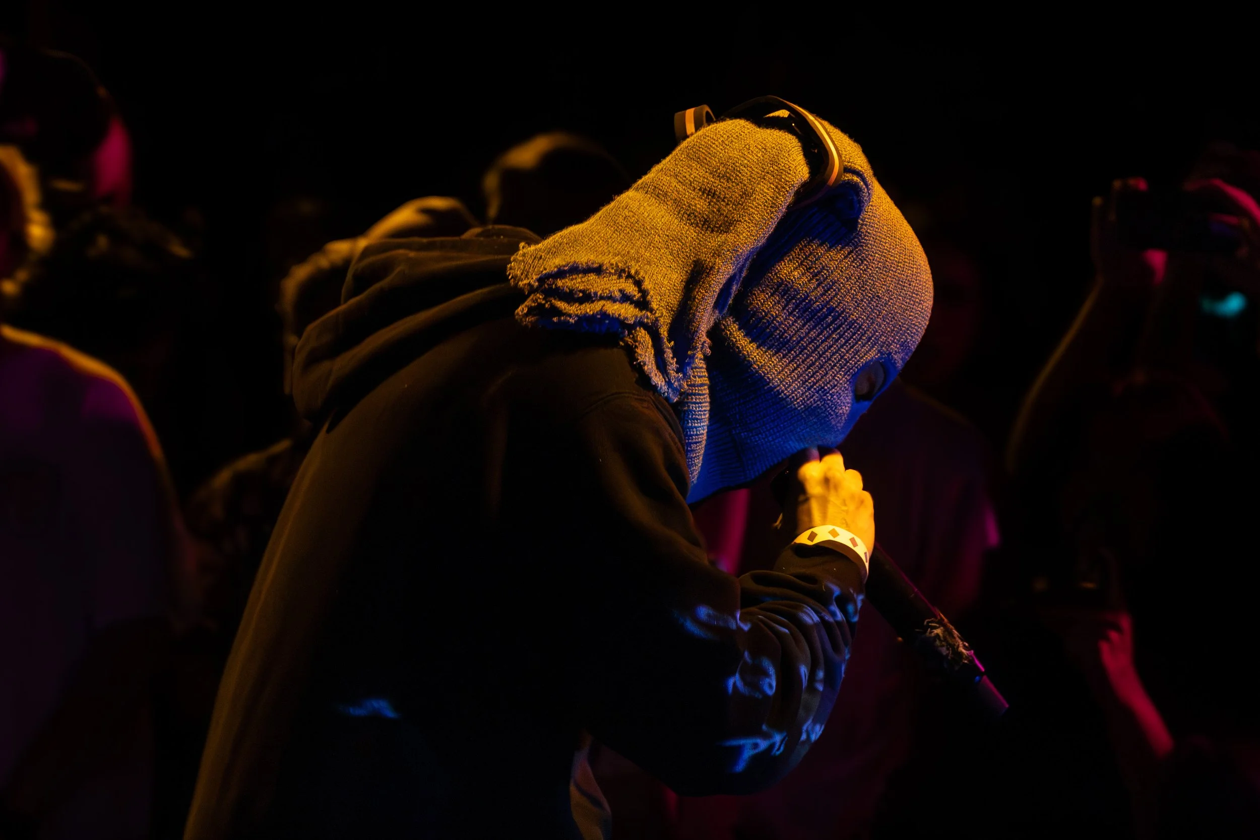 A person wearing a gray knit hoodie with yellow highlights, headphones, and a black jacket, is seen holding a microphone with hands pressed together in front of face during a dark event.