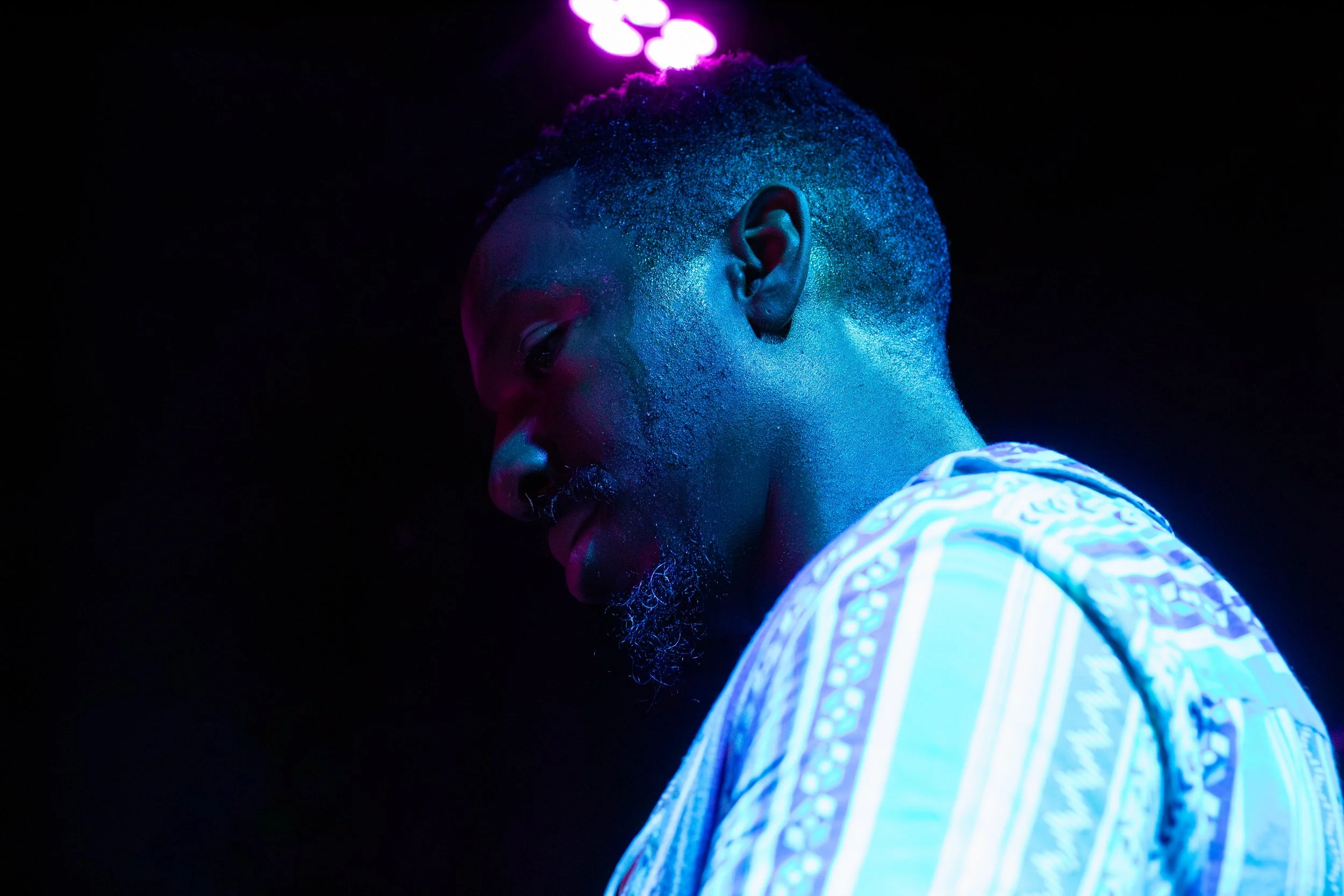 A side profile of a man with short curly hair and a beard, illuminated by colorful stage lighting, wearing a patterned shirt, with a dark background.