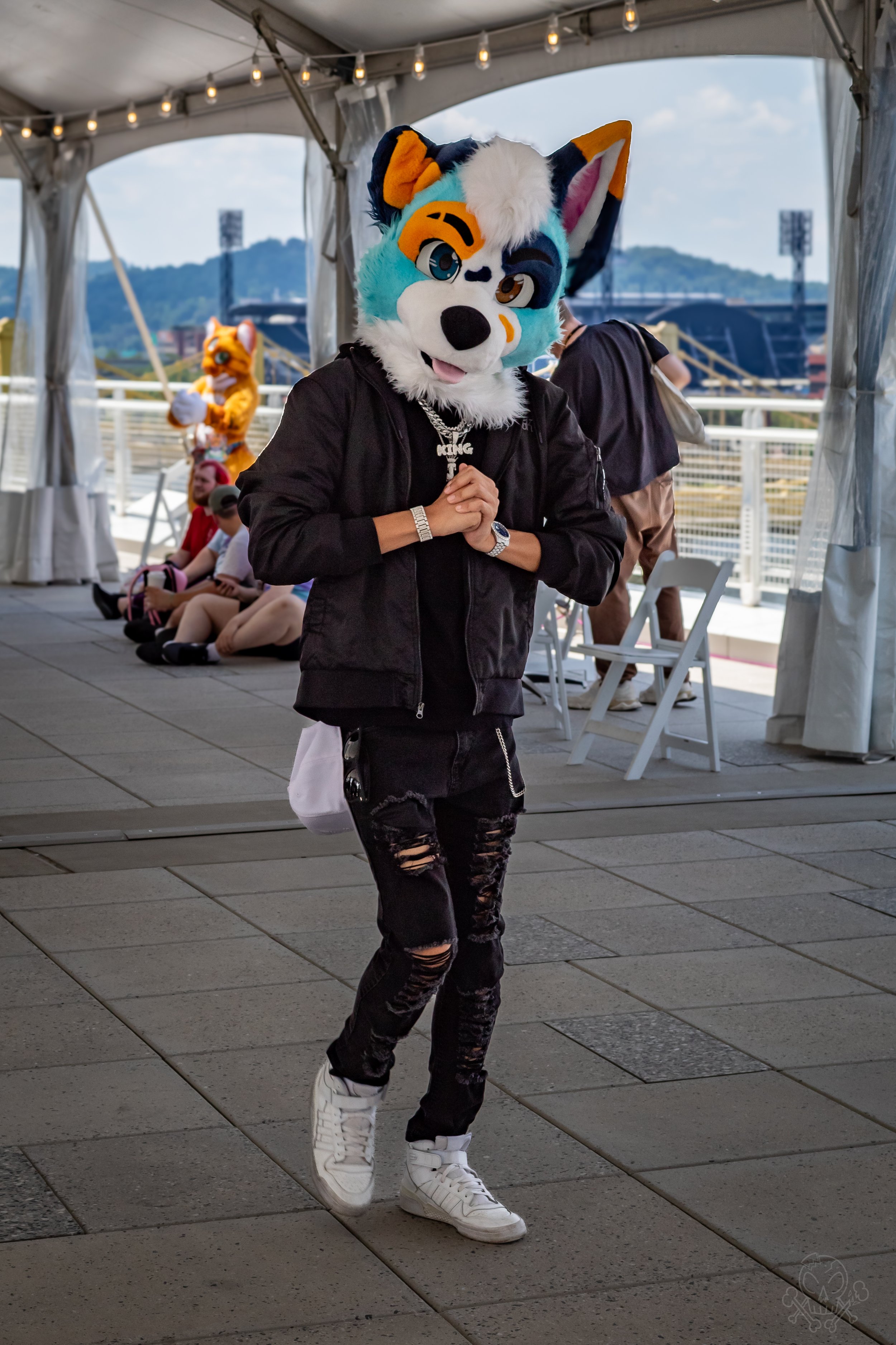 Person in a colorful wolf costume walking on a paved outdoor area, with other people sitting in the background under a tent.