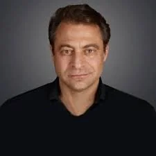 A man with short dark hair and a slight smile, wearing a black shirt against a gray background.