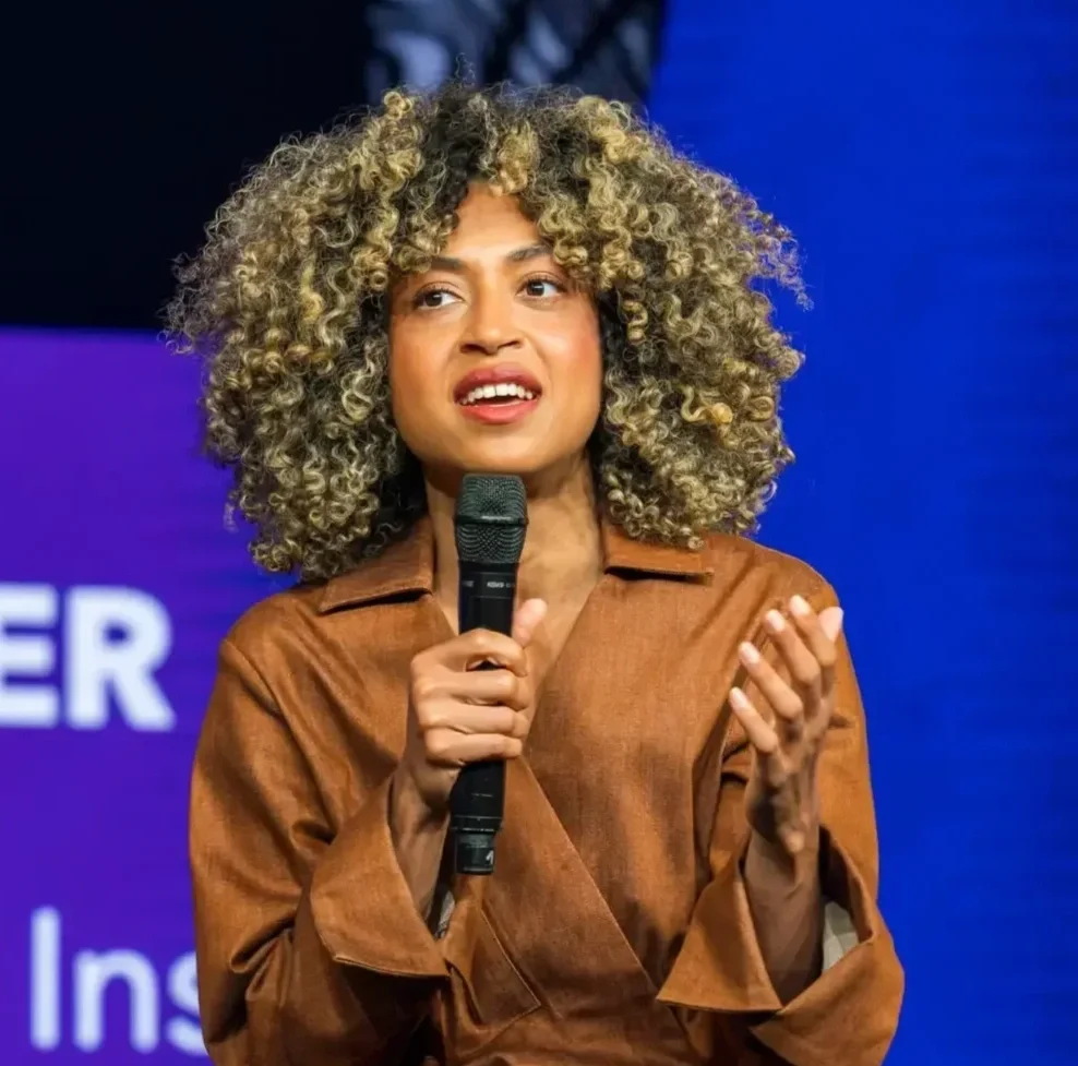 Woman with curly blonde hair speaking into a microphone on stage, wearing a brown jacket.