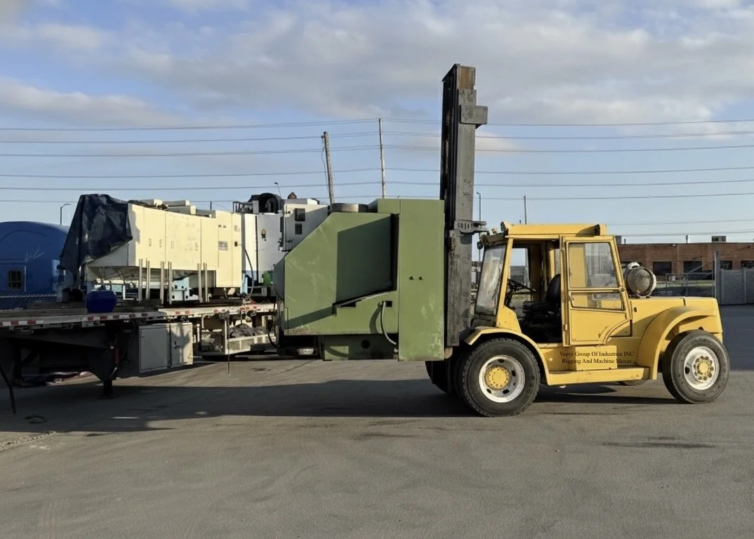 A yellow forklift moving a large machine on a flatbed truck in an outdoor industrial area.