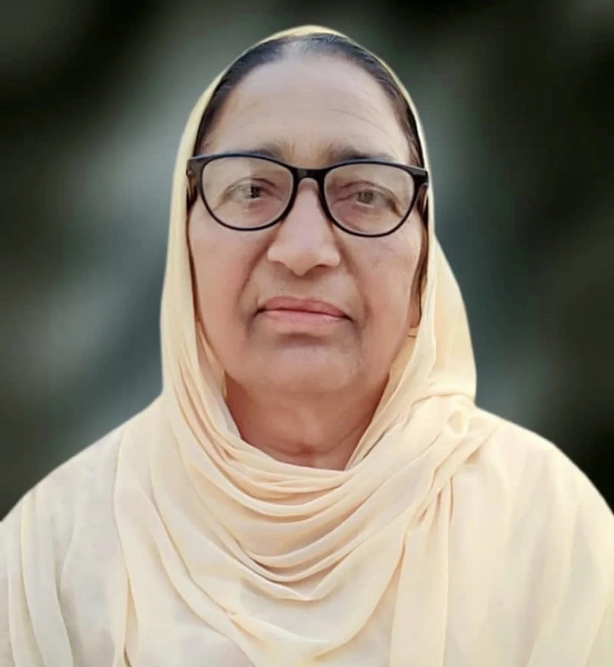 A woman wearing glasses and a light-colored headscarf, looking at the camera against a blurred dark green background.