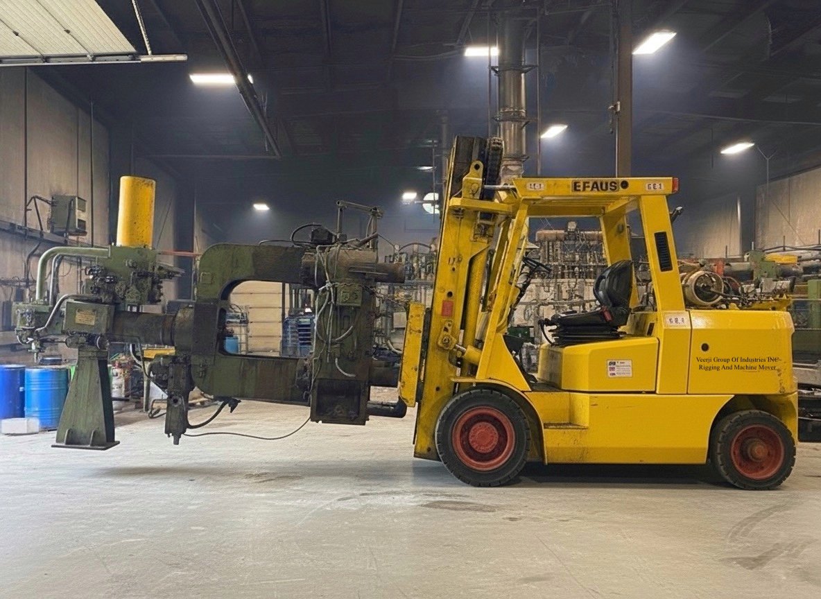 Yellow forklift moving industrial machinery inside a large factory with dim lighting and blue barrels in the background.