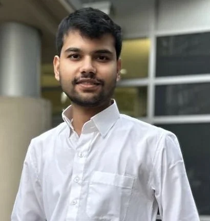 A young man with a beard and short hair, wearing a blue patterned button-up shirt and black pants, standing outdoors in a garden with trees, shrubs, and a house in the background.