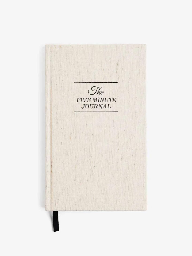 A beige fabric-covered journal titled 'The Five Minute Journal' with a black ribbon bookmark.