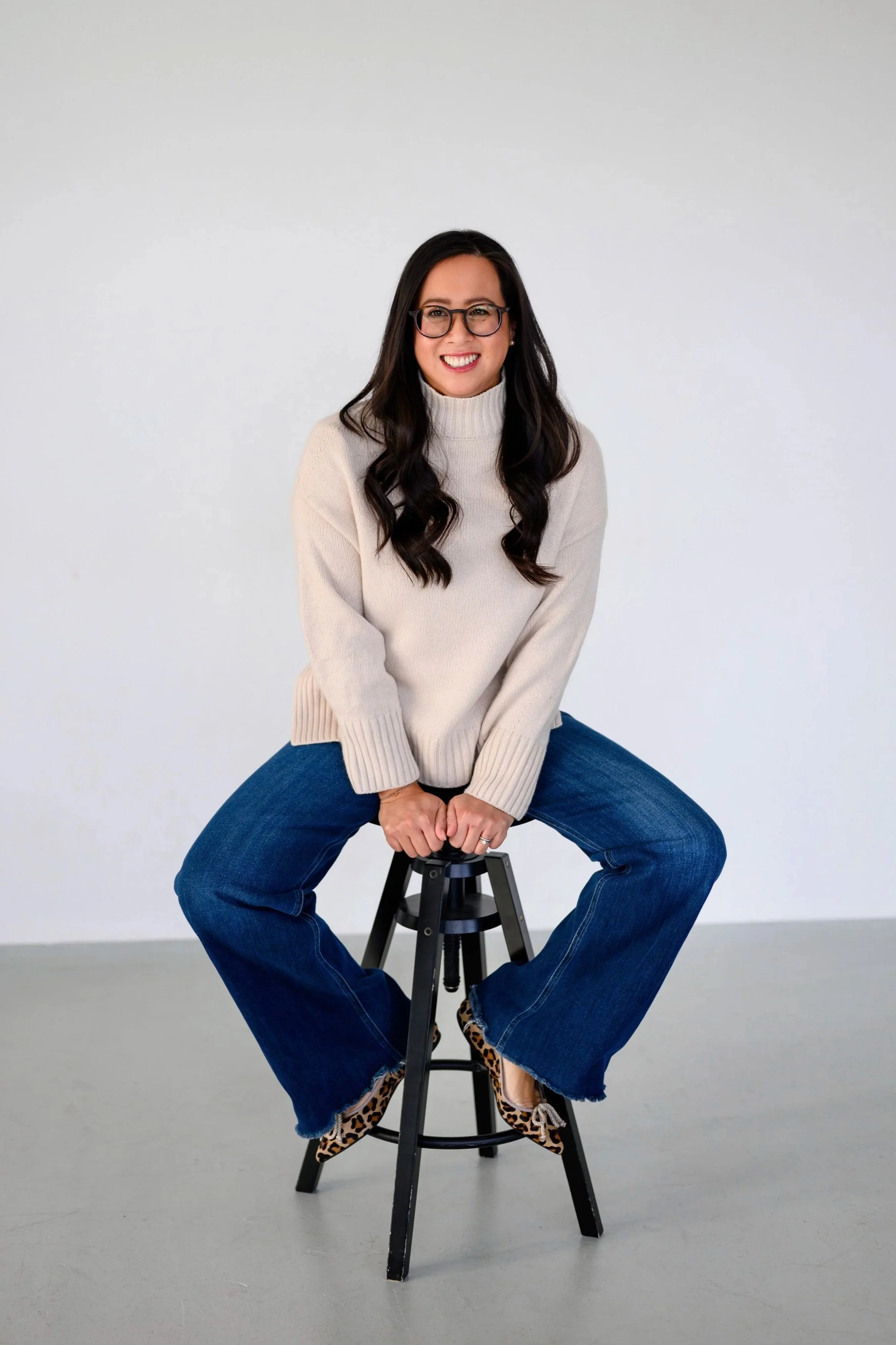 A woman sitting on a black stool in front of a plain white background, wearing glasses, a cream-colored sweater, and blue jeans with leopard print shoes, smiling at the camera.