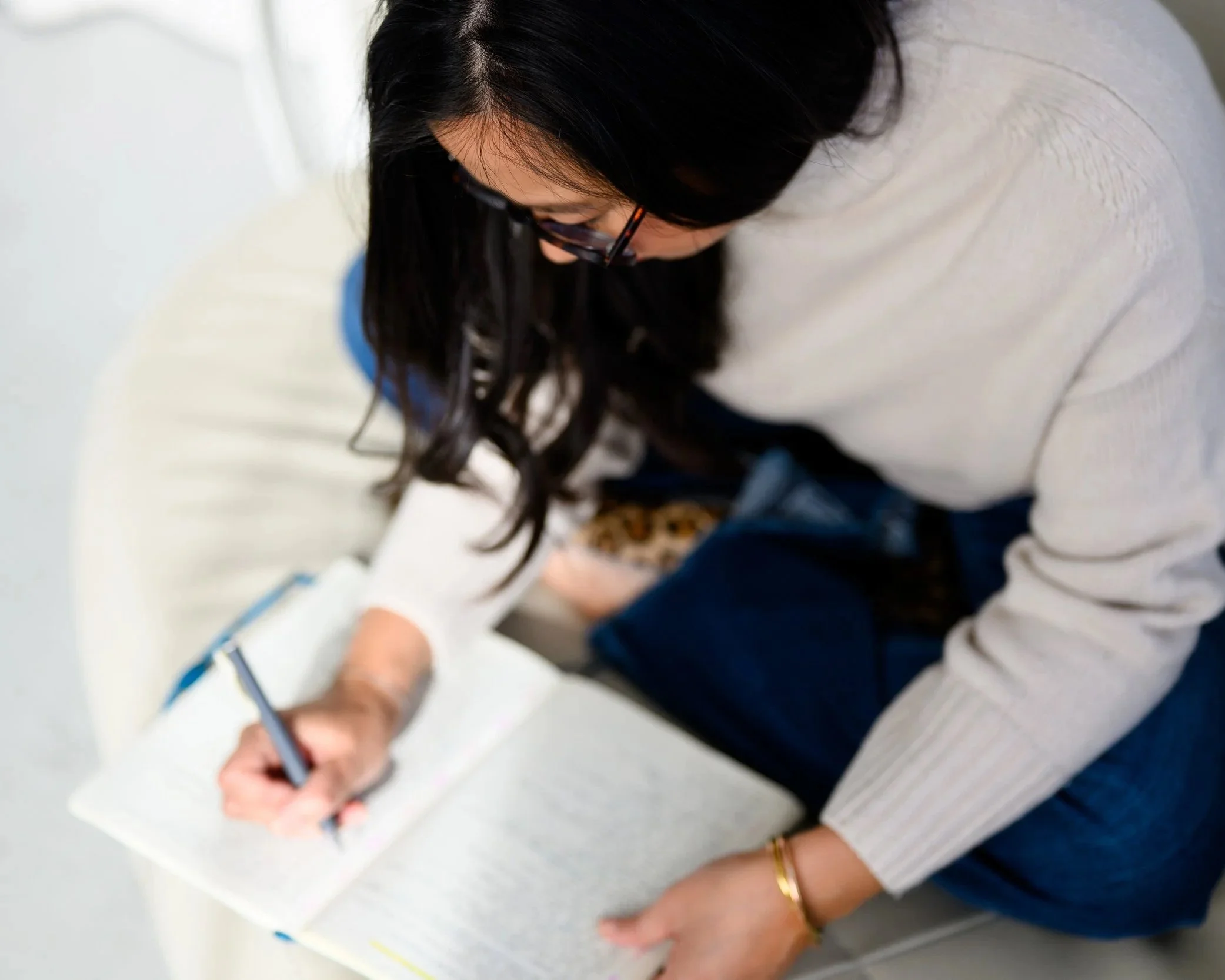 A woman with long dark hair and glasses, sitting on a light-colored surface, writing in a notebook with a pen, dressed in a light sweater and jeans.