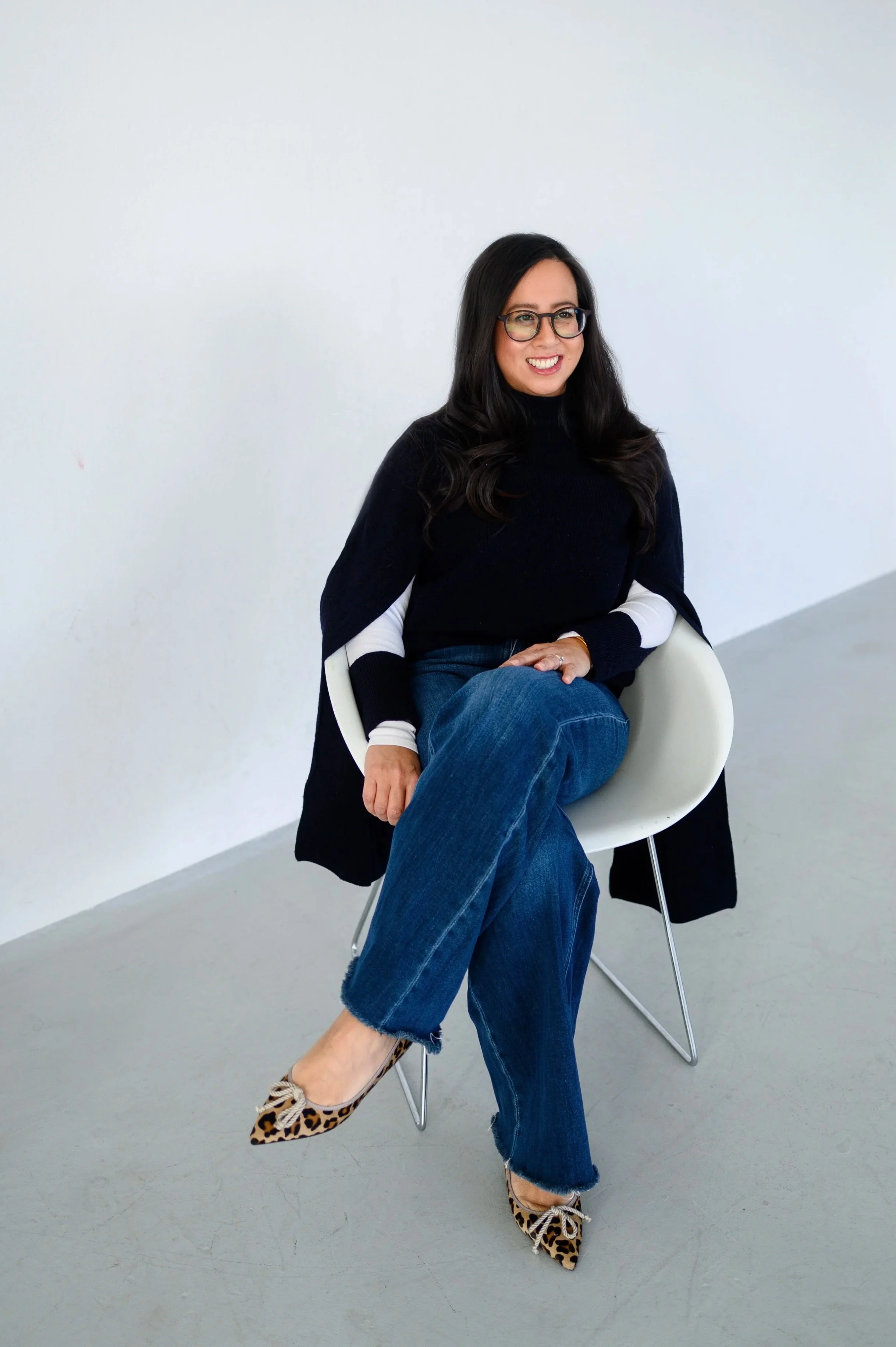 Woman with long dark hair and glasses sitting on a modern white chair against a plain white wall, smiling, wearing a black and white sweater, blue jeans, and leopard print shoes.