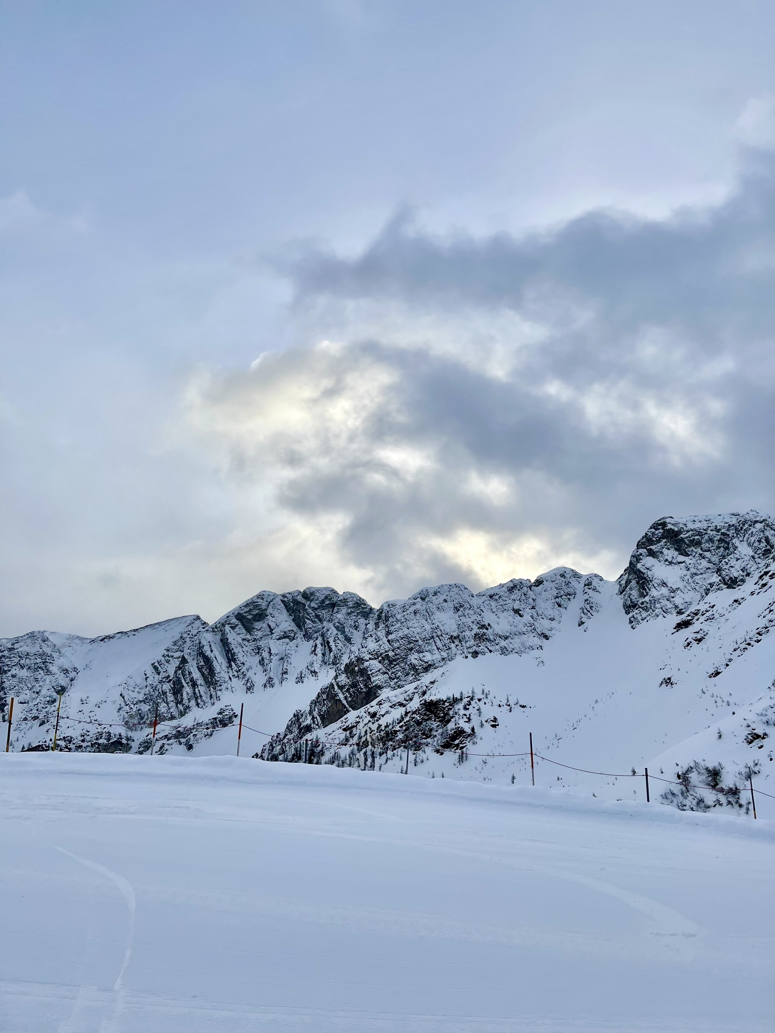 Snow-covered mountain landscape with cloudy sky, ski slope, and orange safety poles.