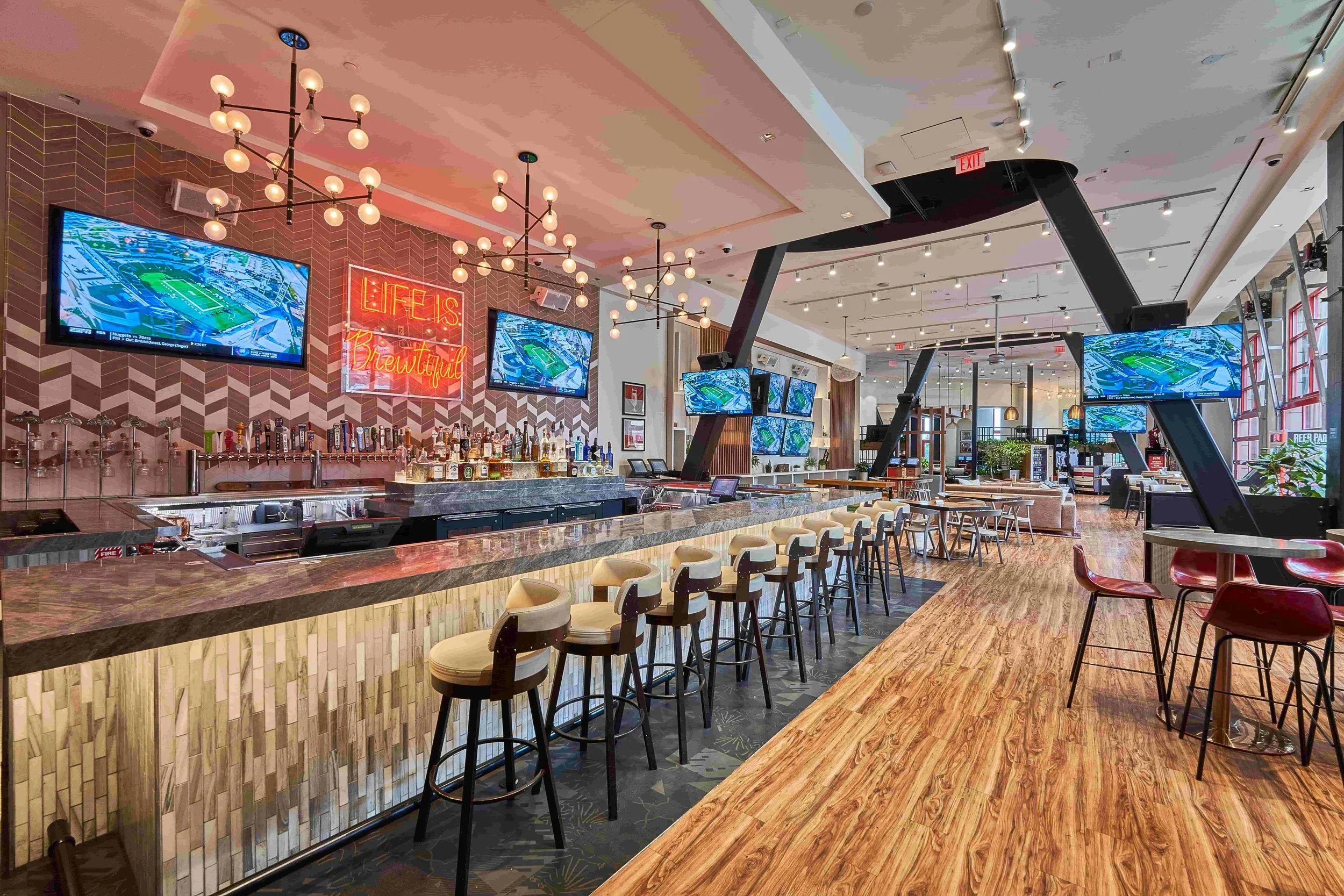 Interior of a modern sports bar with multiple TVs showing a stadium, a bar counter with stools, and a cozy seating area with tables and chairs. Stylish lighting fixtures and decorative elements are visible.