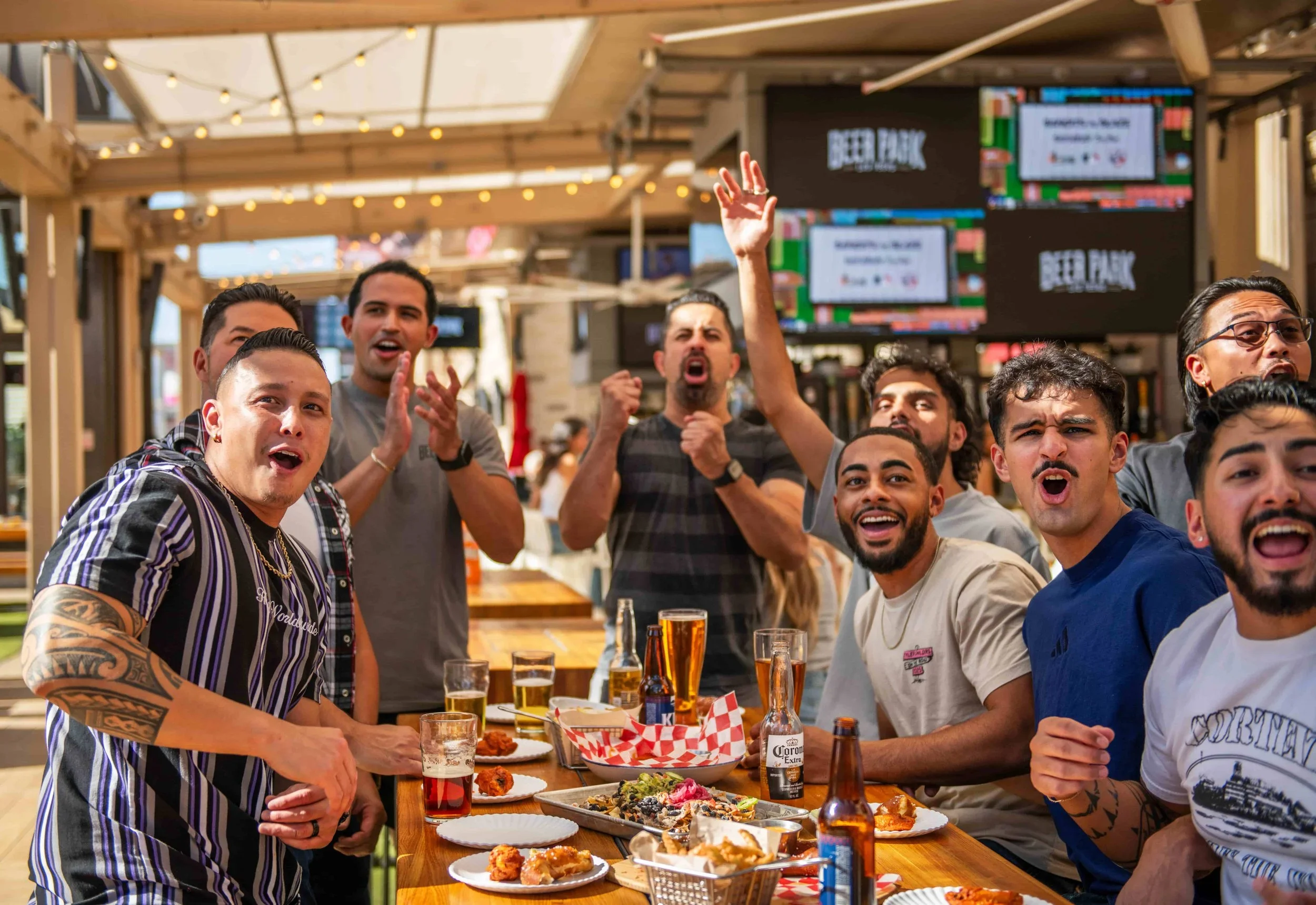 Group of friends celebrating at a restaurant or bar, with food and drinks on the table, cheering, smiling, and raising their hands in excitement.