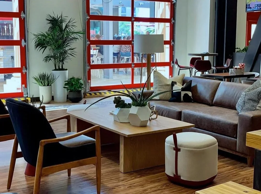 Modern cafe lounge with chairs, a leather sofa, and a coffee table decorated with potted plants. Large glass windows show people outside at tables.