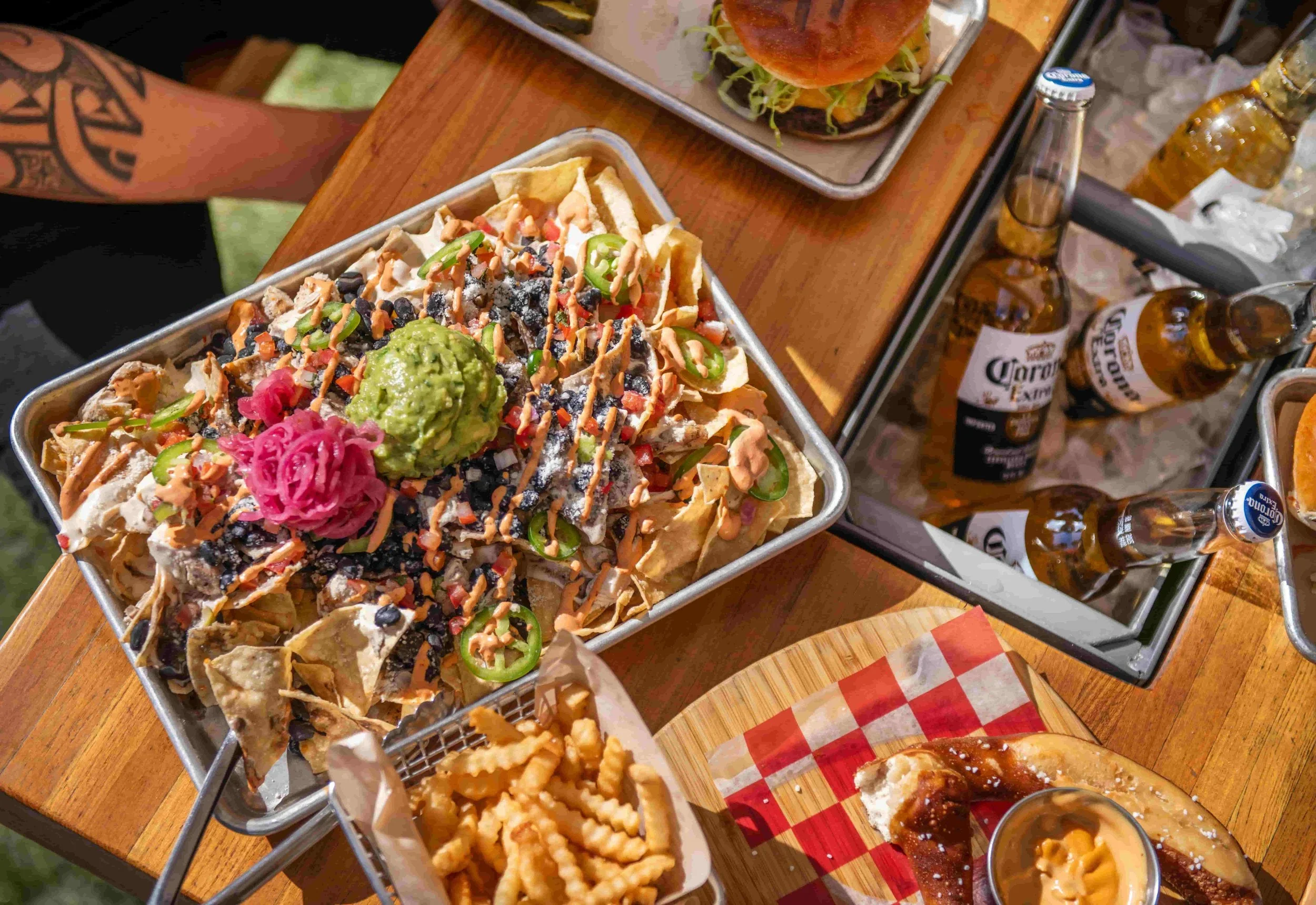 A tray of loaded nachos topped with guacamole, pink pickled onions, jalapeños, black beans, shredded cheese, and various sauces on a picnic table. Nearby are bottles of Corona beer, a basket of French fries, a slice of pizza with toppings, and a glass of beer. There is a hamburger with lettuce, tomato, and bun on a tray in the background.