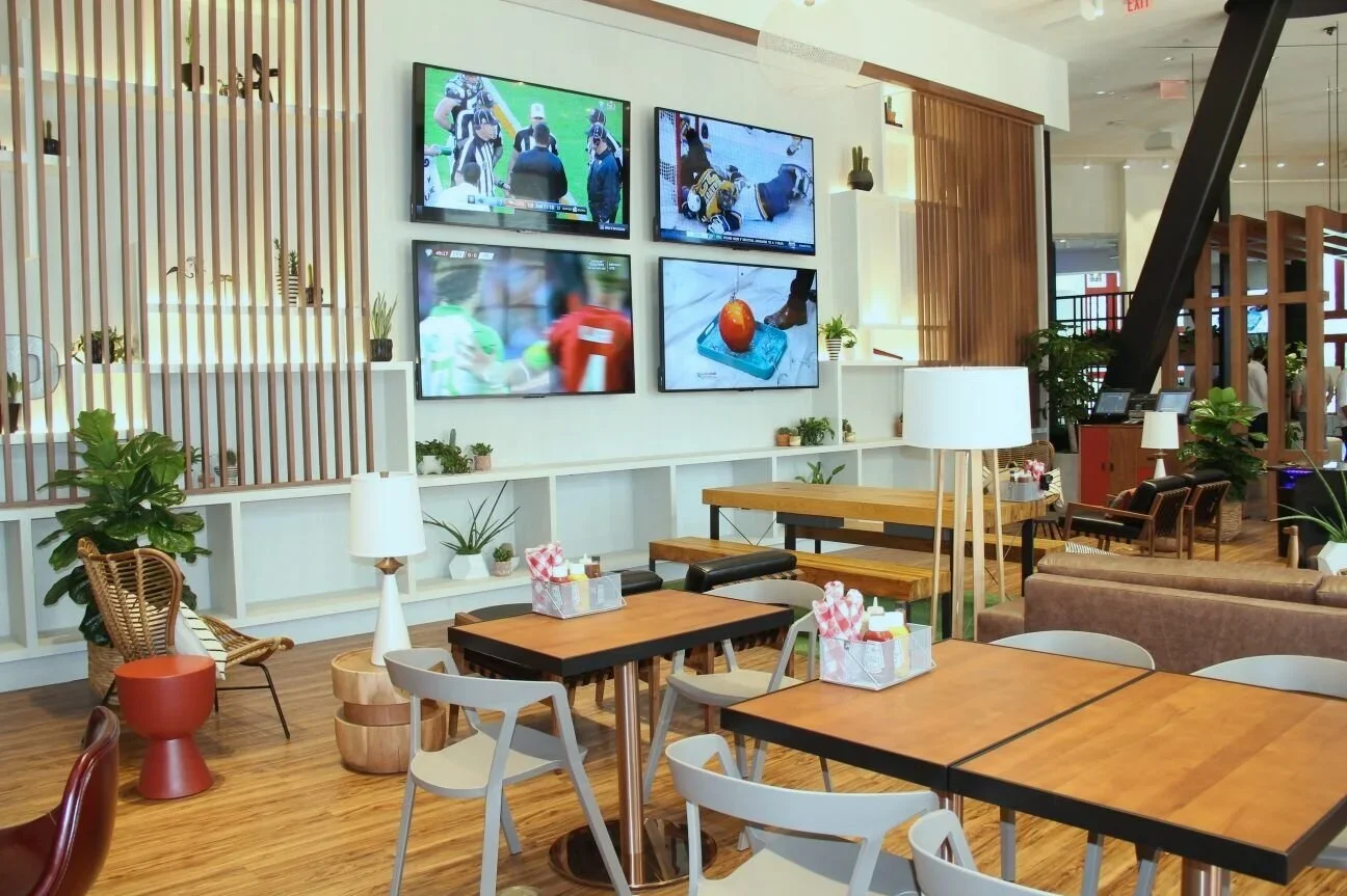 Interior of a modern cafe or lounge area with wooden tables and chairs, potted plants, white lamps, and four mounted television screens showing a football game, with other patrons visible in the background.