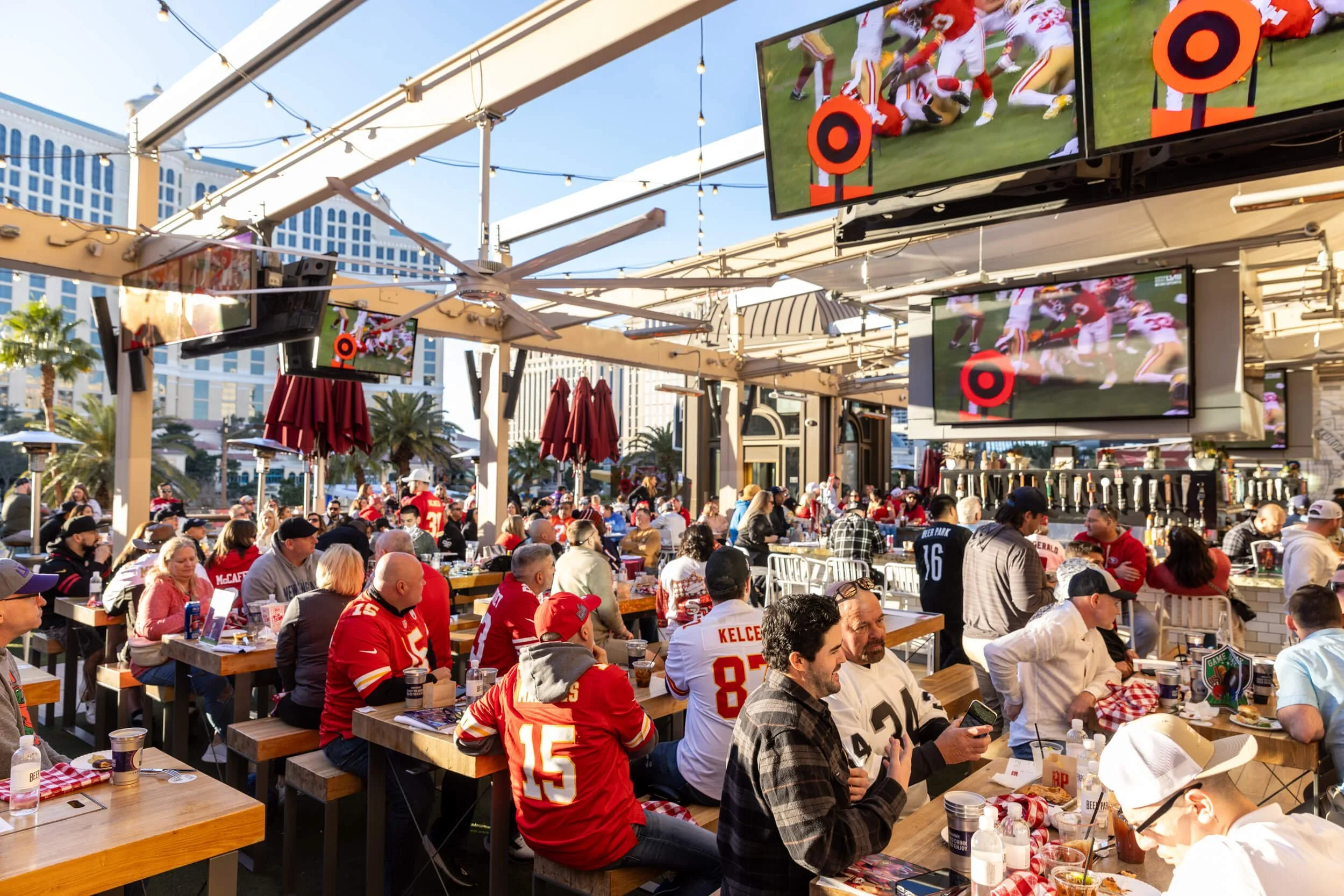 Crowd of people watching a football game on large outdoor screens at a sports bar with string lights and high-rise buildings in background.
