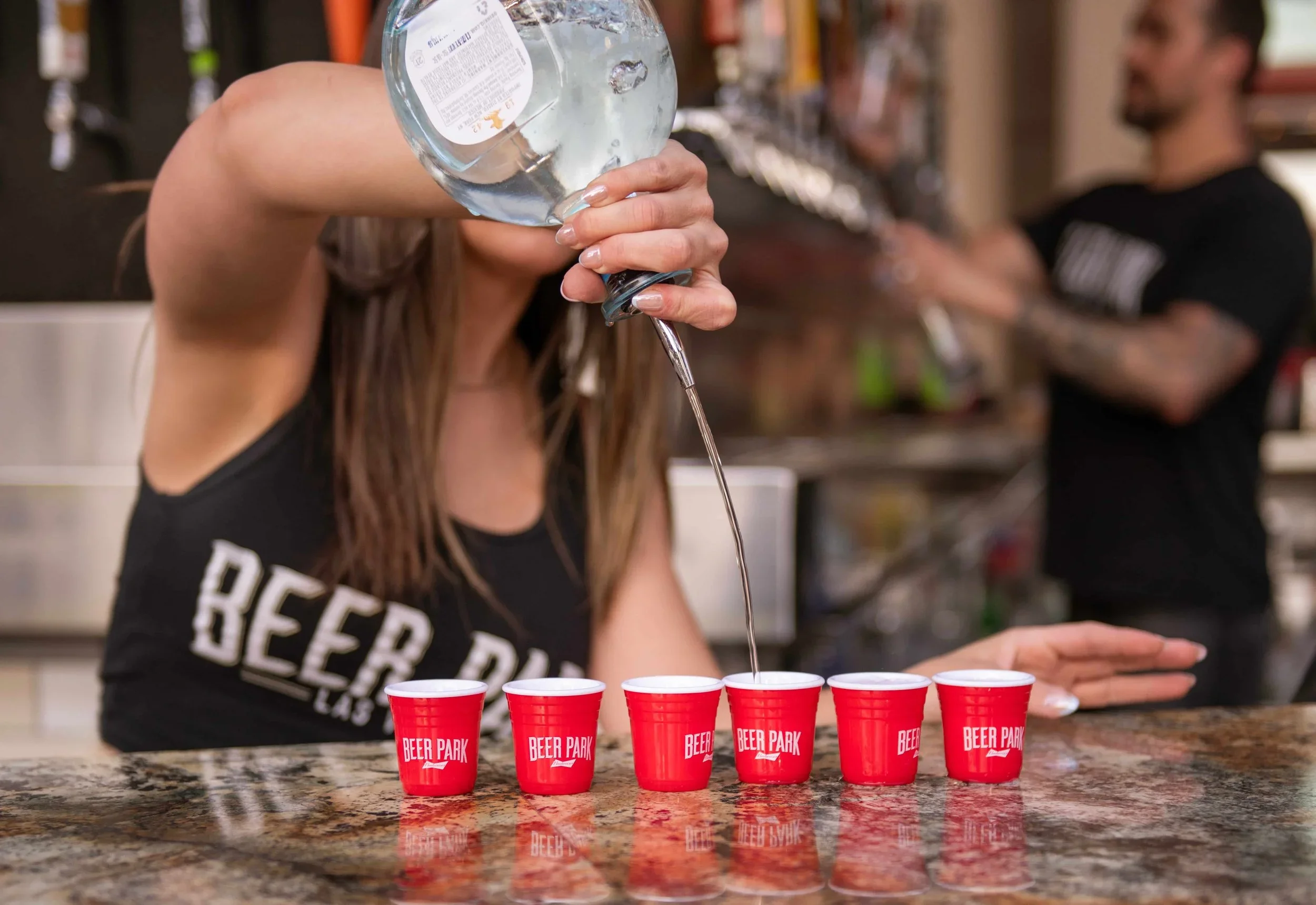 A woman is pouring clear liquid from a plastic bottle into red cups on a bar counter with 'Beer Park' written on them, while a man stands in the background.