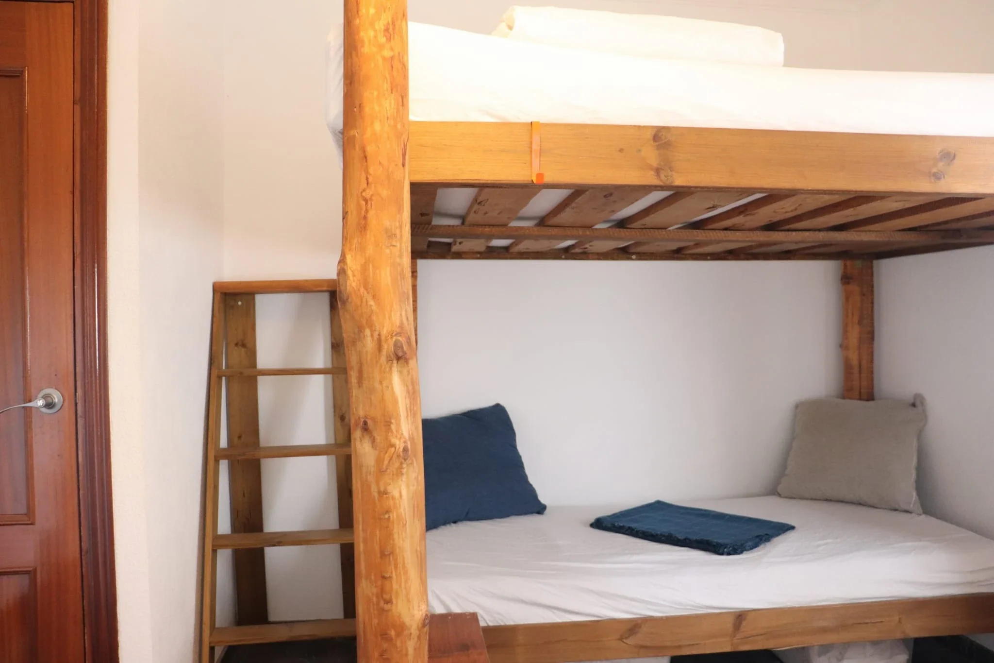 Wooden bunk bed with white bedding, gray pillow, blue pillow, and blue blanket in a room with white wall and wooden door.