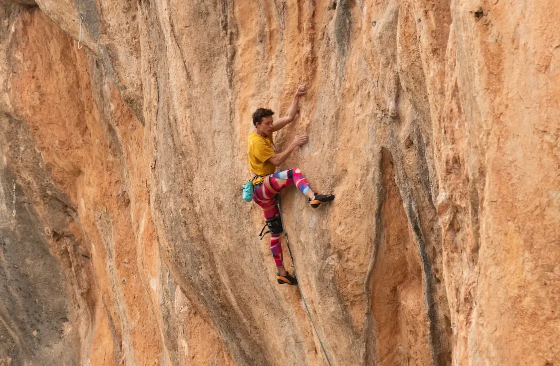 A Climbing guide wearing colorful climbing pants is rock climbing on a tall, rugged orange-brown cliff face.