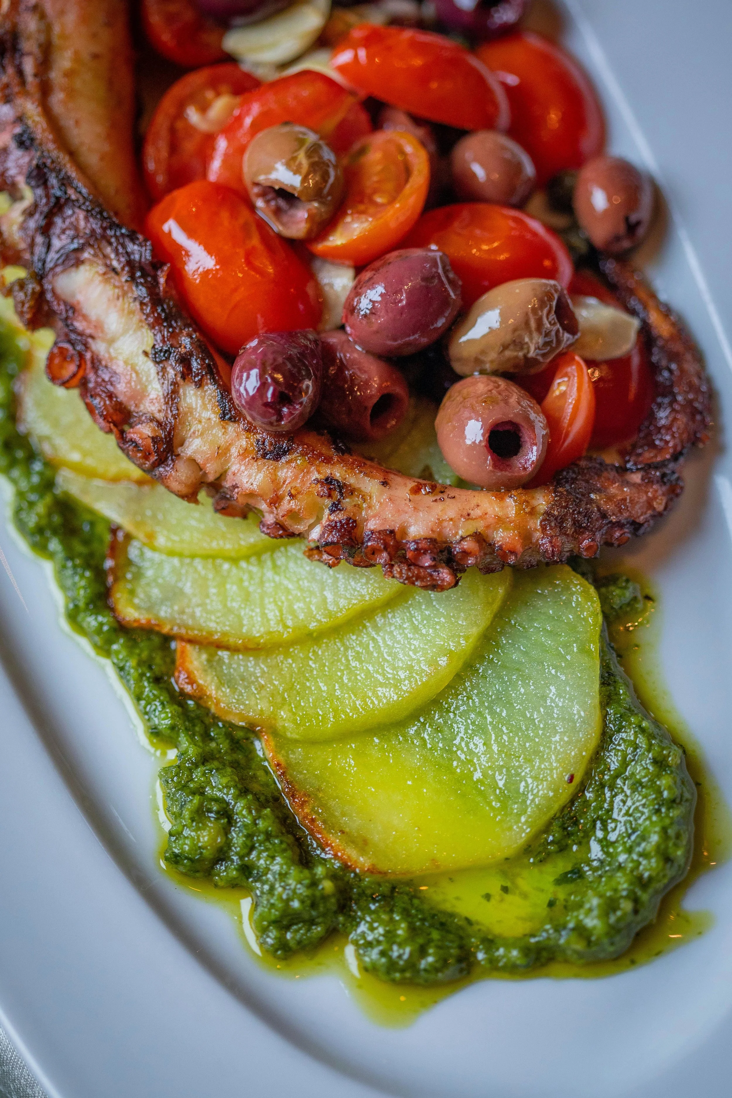 Grilled octopus tentacle served with cherry tomatoes, olives, slices of zucchini, and green sauce on a white plate.
