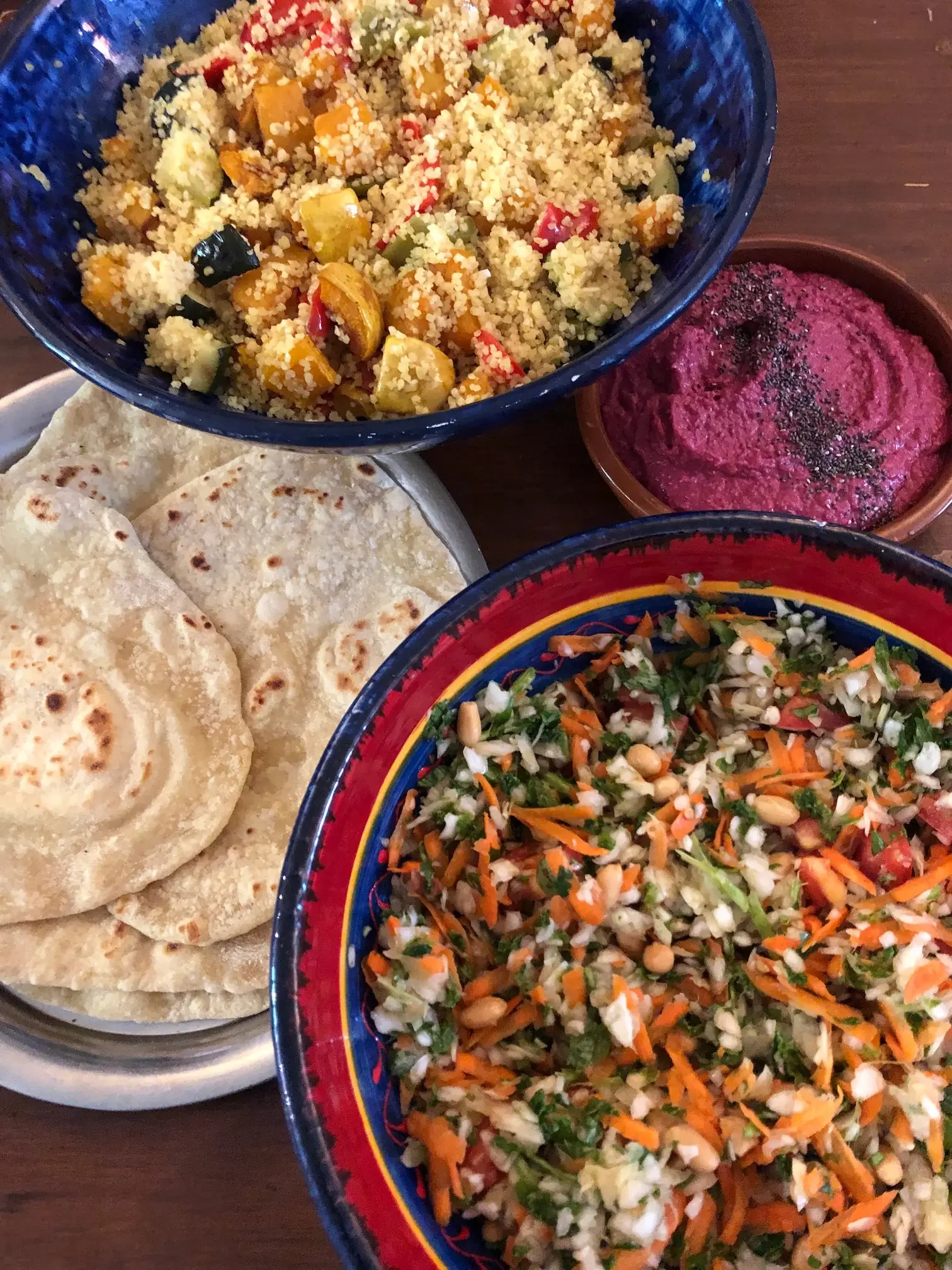 A variety of colorful Mexican dishes including a bowl of couscous salad with vegetables, a plate of warm tortillas, a bowl of mashed beet dip topped with black pepper, and a bowl of fresh vegetable salad with shredded carrots, cabbage, and herbs.