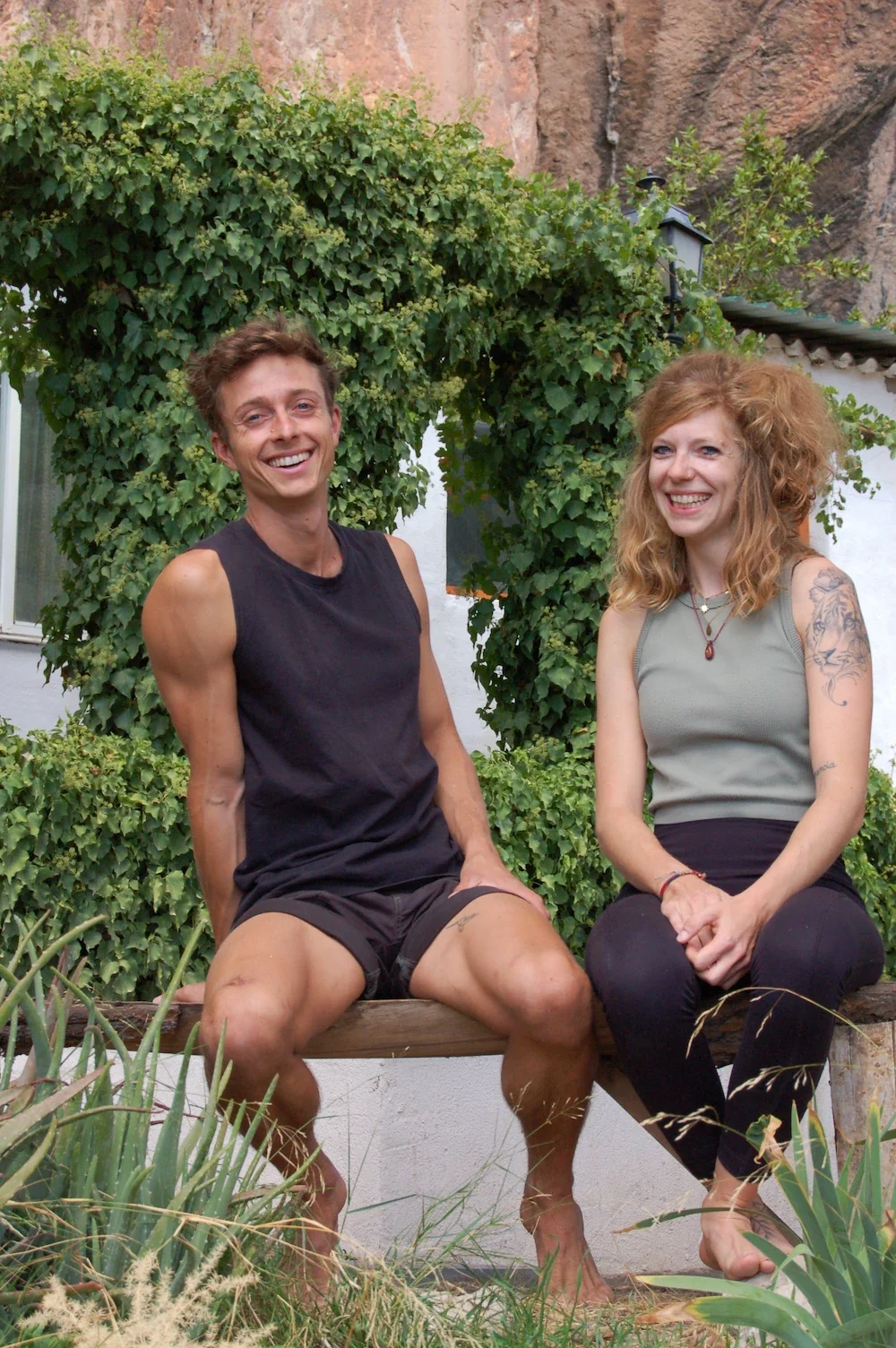 A Yoga teacher and a Climbing instructor smilling and sitting outdoors on a wooden bench in front of greenery, with a rock wall in the background.