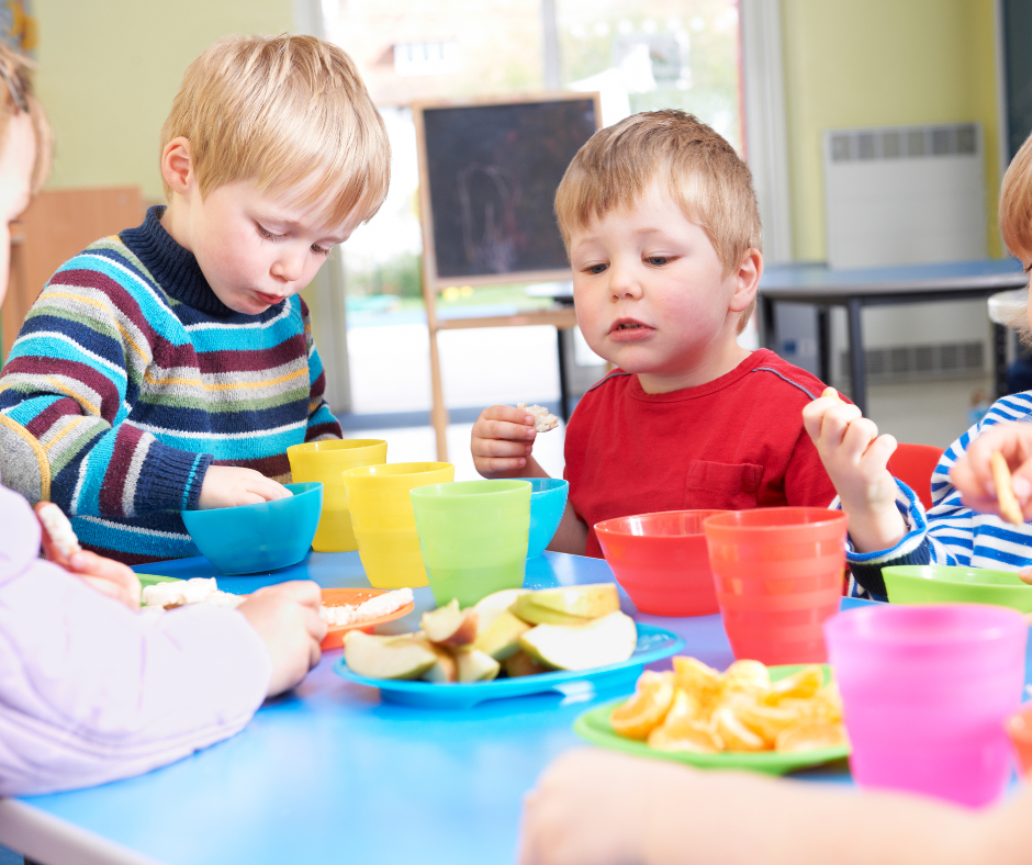 Children sitting at a table with colorful bowls and plates of snacks, including cut fruit and chips, in a classroom setting.