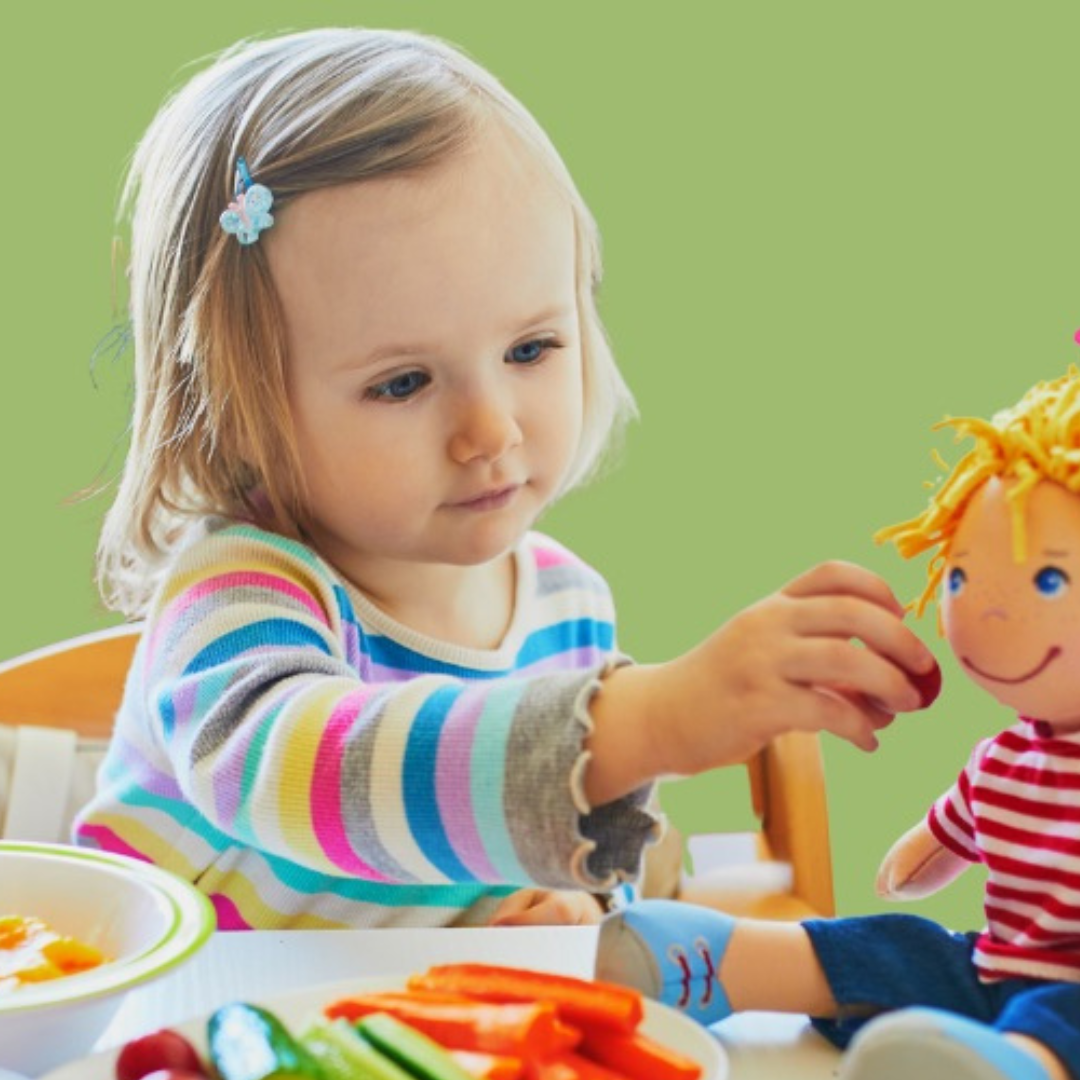A young girl with blonde hair and a butterfly hair clip, wearing a colorful striped long sleeve shirt, playing with a rag doll at a table with food and vegetables, against a green background.