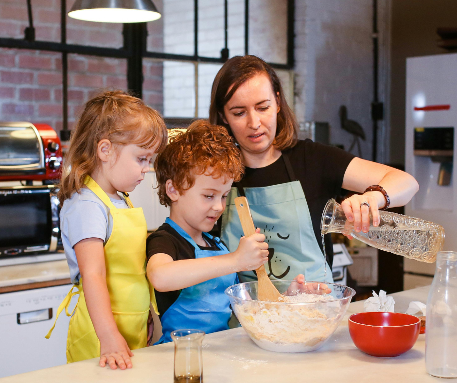 A woman and two children baking together in a kitchen, with the woman pouring water and the children stirring a large mixing bowl.