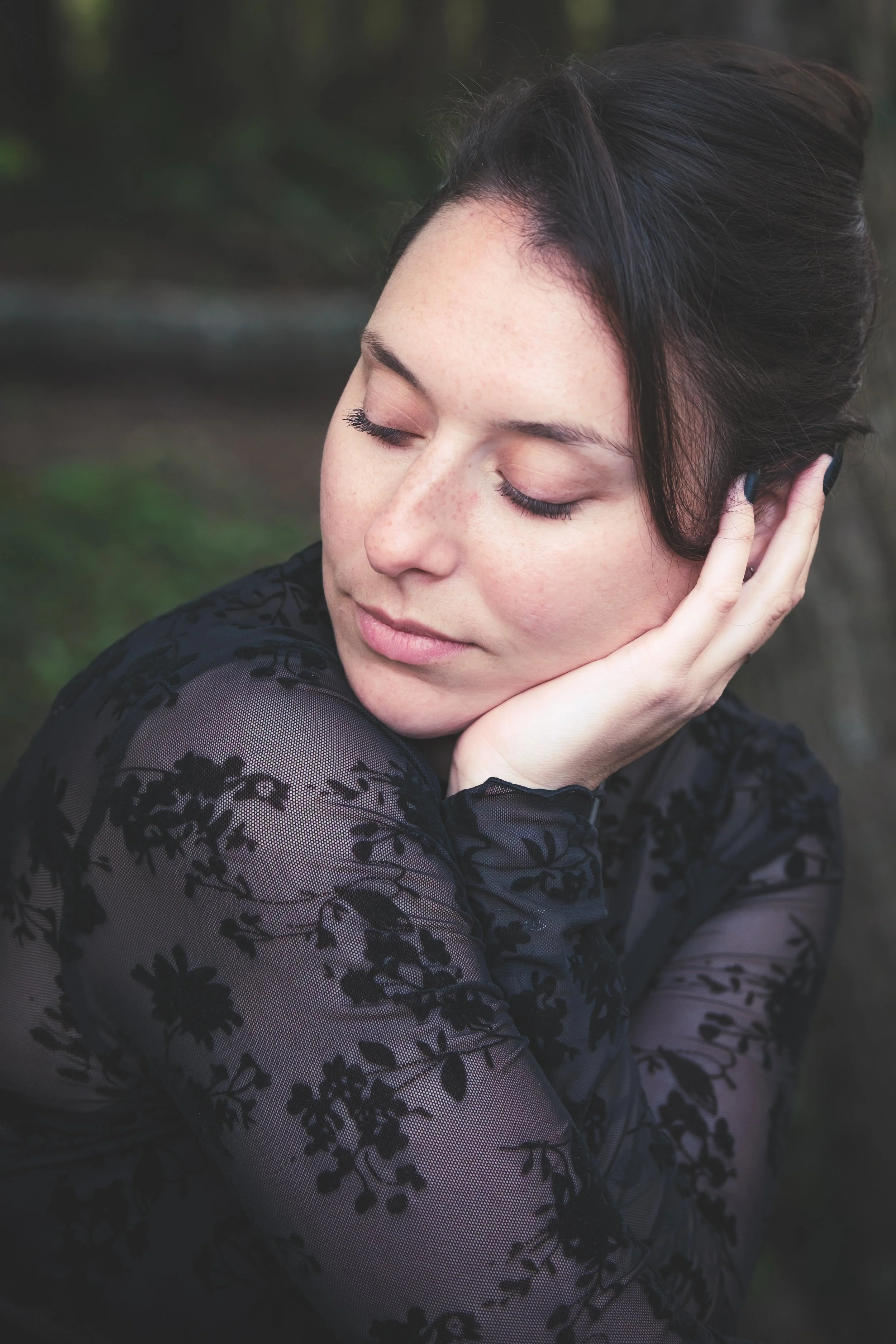 Jeune femme aux yeux fermés, portant un vêtement en dentelle noire avec un motif floral, regardant vers le bas, dans un environnement naturel.