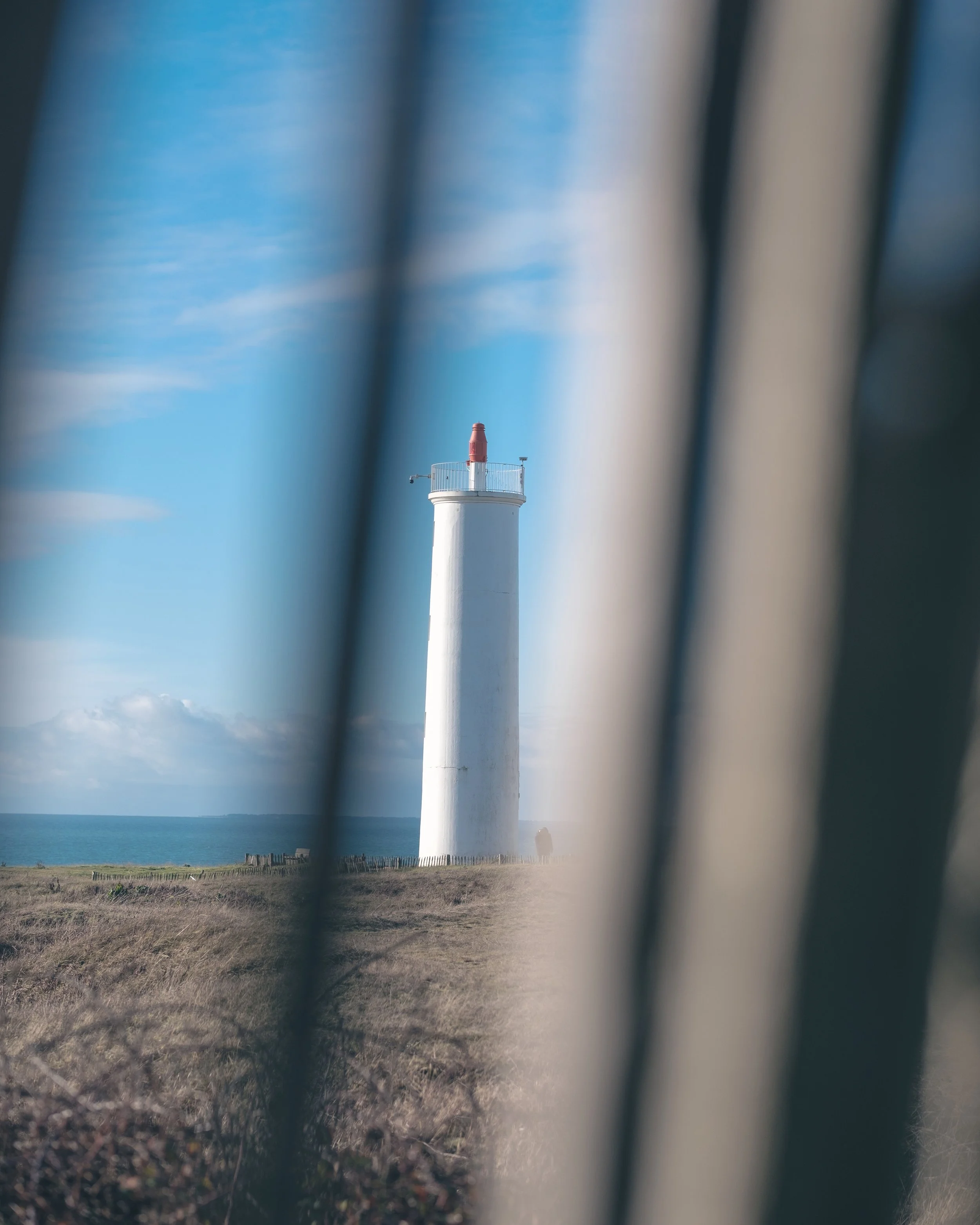Phare blanc avec une rambarde et une lanterne rouge au sommet, vu à travers des barres noires et une clôture en bois sur un fond de ciel bleu et mer.