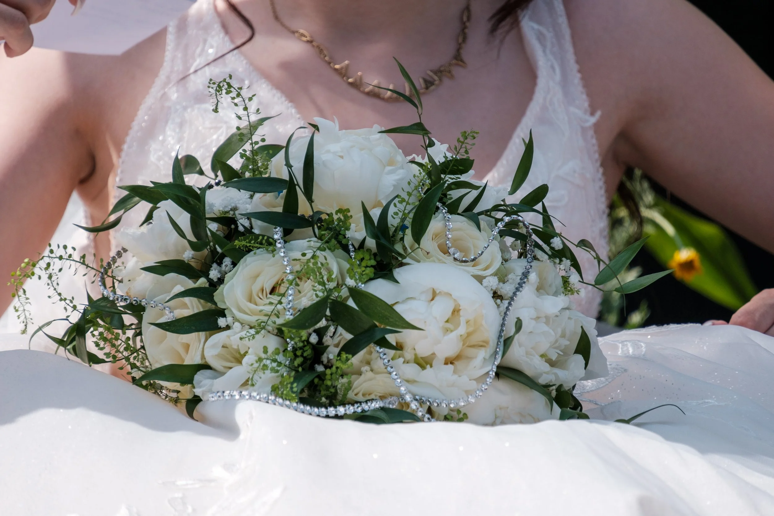 Bouquet de mariage composé de roses blanches, décoré avec des bijoux en perles et des feuilles vertes, porté par une mariée en robe blanche avec un collier or et un corsage en dentelle.