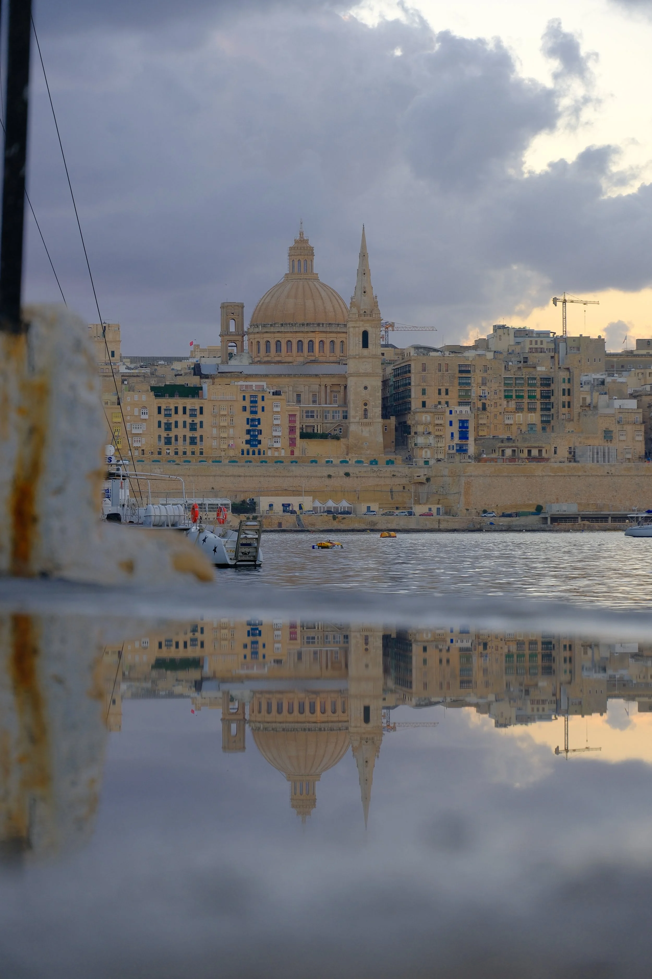 Vue du port de La Valette avec une reflection dans l'eau, montrant une grande coupole et une église sur l'horizon, sous un ciel nuageux.