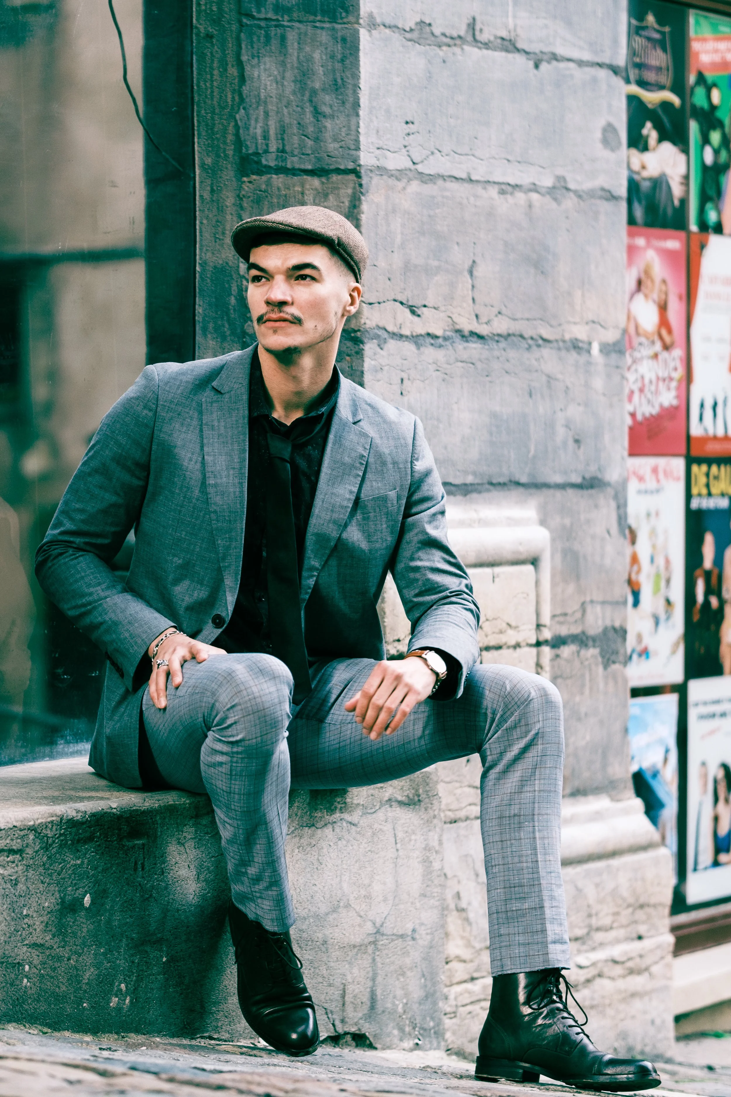 Un homme assis sur un rebord de pierre, portant un costume gris à carreaux, une chemise noire, des bottines en cuir noir, une montre et un chapeau gris, avec un mur en pierre en arrière-plan et des affiches colorées à droite.