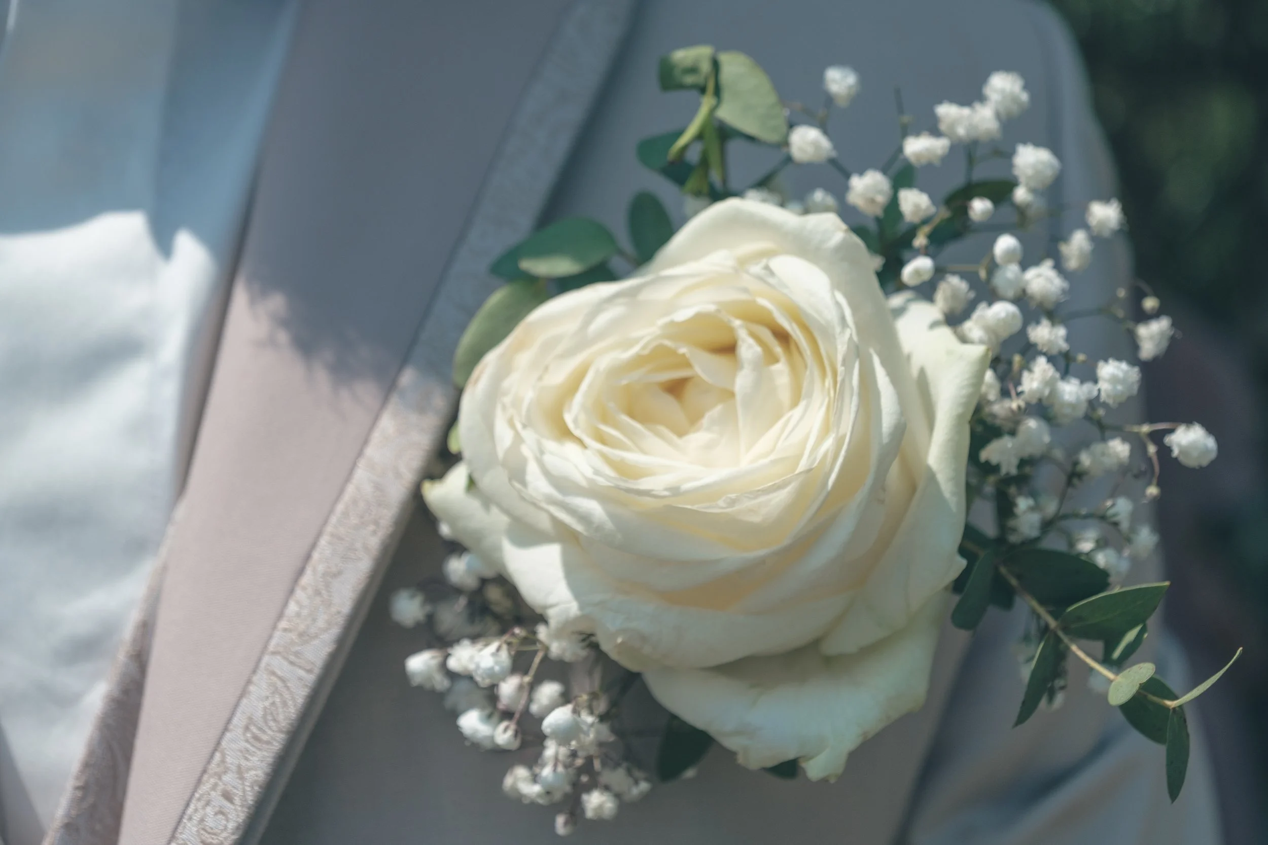 Une composition florale avec une rose blanche entourée de petites fleurs blanches et de feuillage vert, posée sur une surface grise.